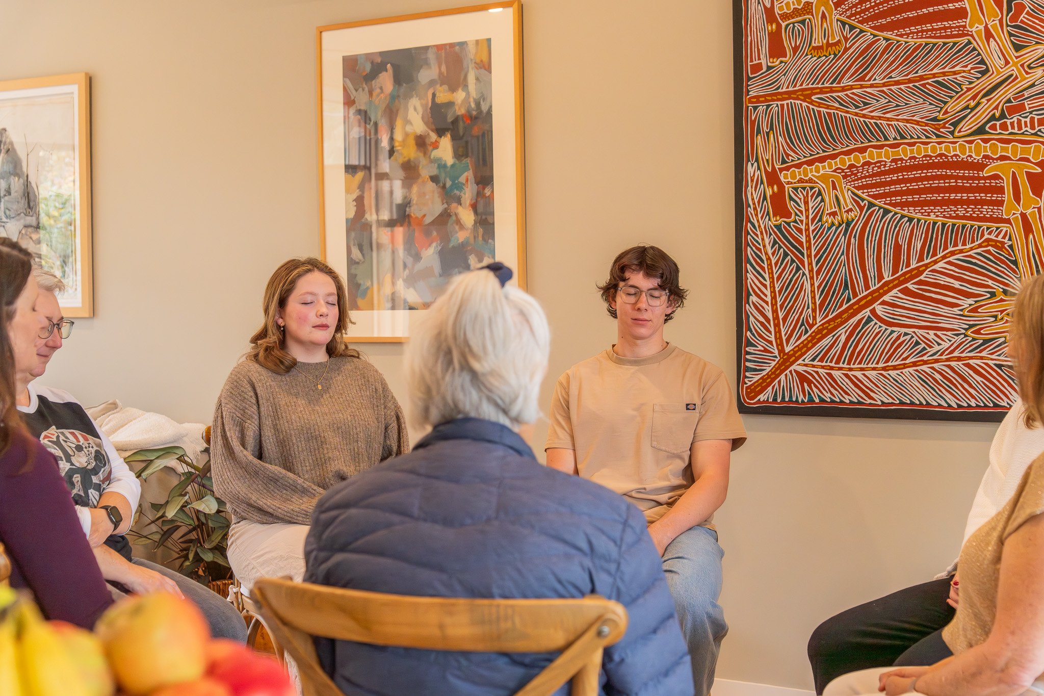People sitting in a circle with eyes closed in an art gallery or museum that features abstract paintings on the wall.