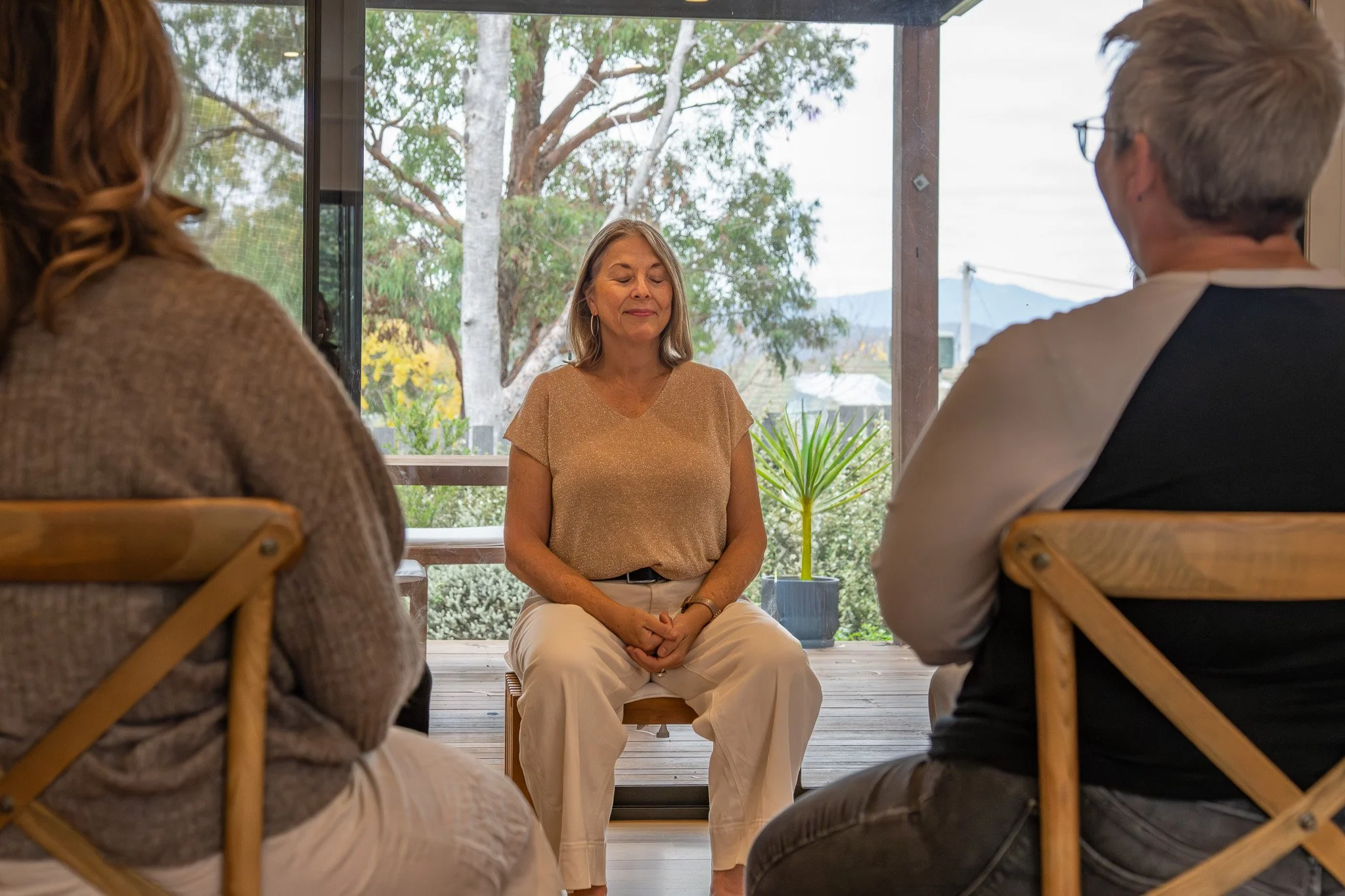 A woman sitting with eyes closed in front of a window, surrounded by two people with their back to the camera, in a room with wooden flooring and plants outside.