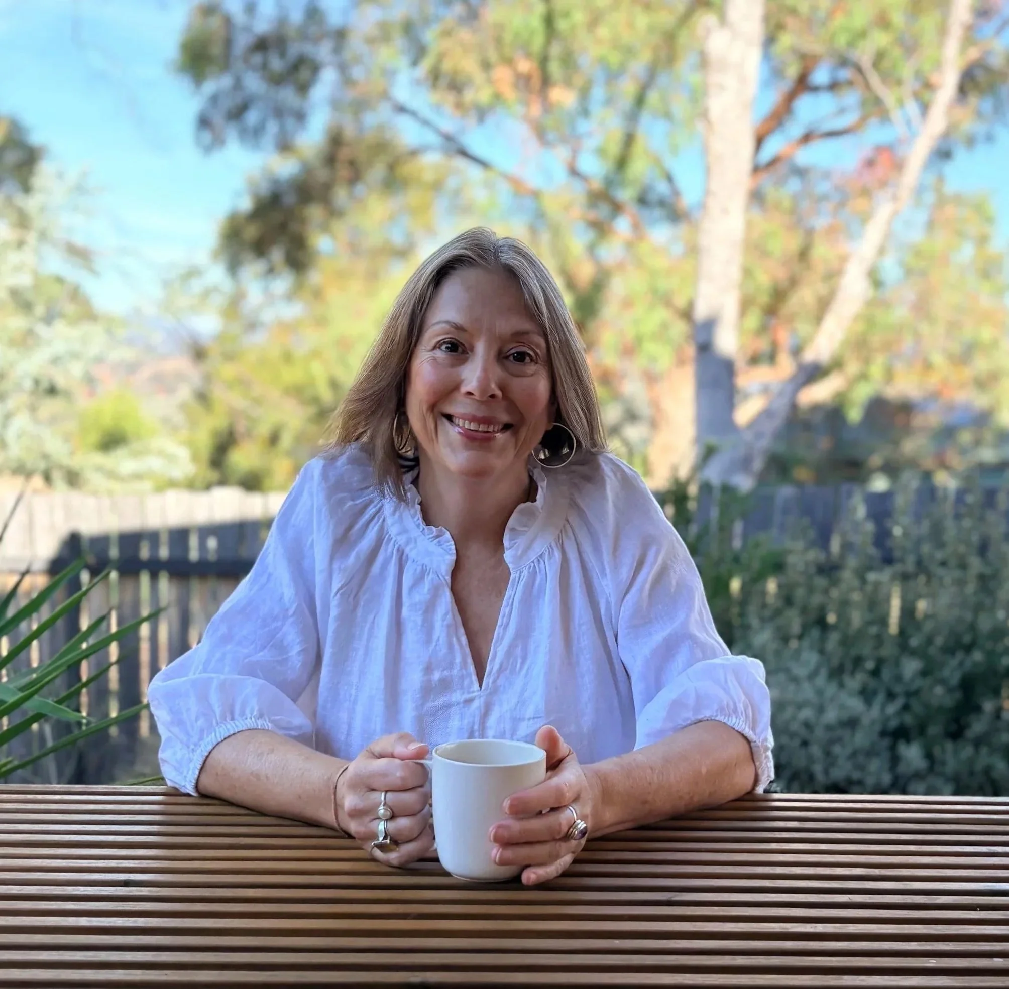 A smiling woman with light brown hair and hoop earrings holds a white mug with both hands, sitting outdoors at a wooden table with a blurred background of trees and sky.