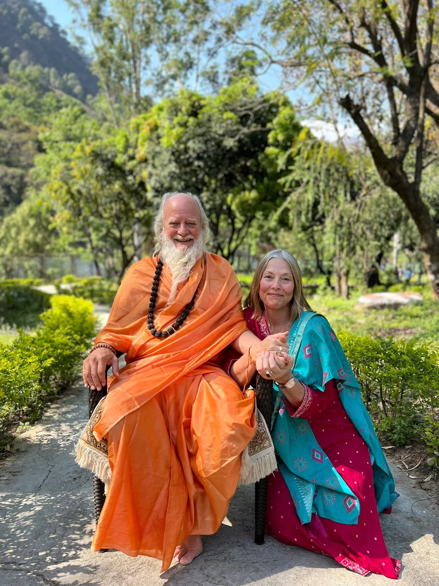 A man and woman sitting together outdoors in a garden with green trees and mountains in the background. The man, with a white beard, is dressed in traditional orange robes with a black beaded necklace, and the woman is wearing a blue and pink traditional dress, holding hands with the man and smiling.