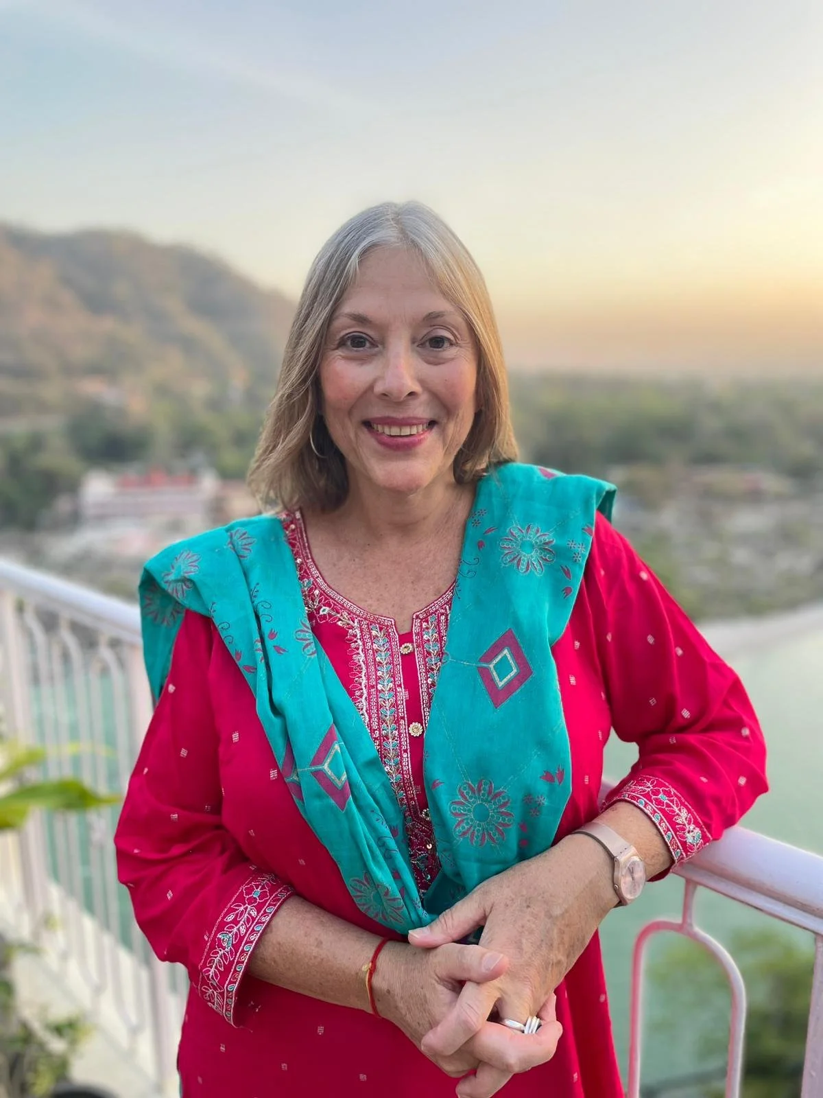 A smiling woman with blonde hair standing outdoors on a balcony during sunset, wearing a pink traditional Indian outfit with a blue embroidered shawl, and a mountain landscape in the background.
