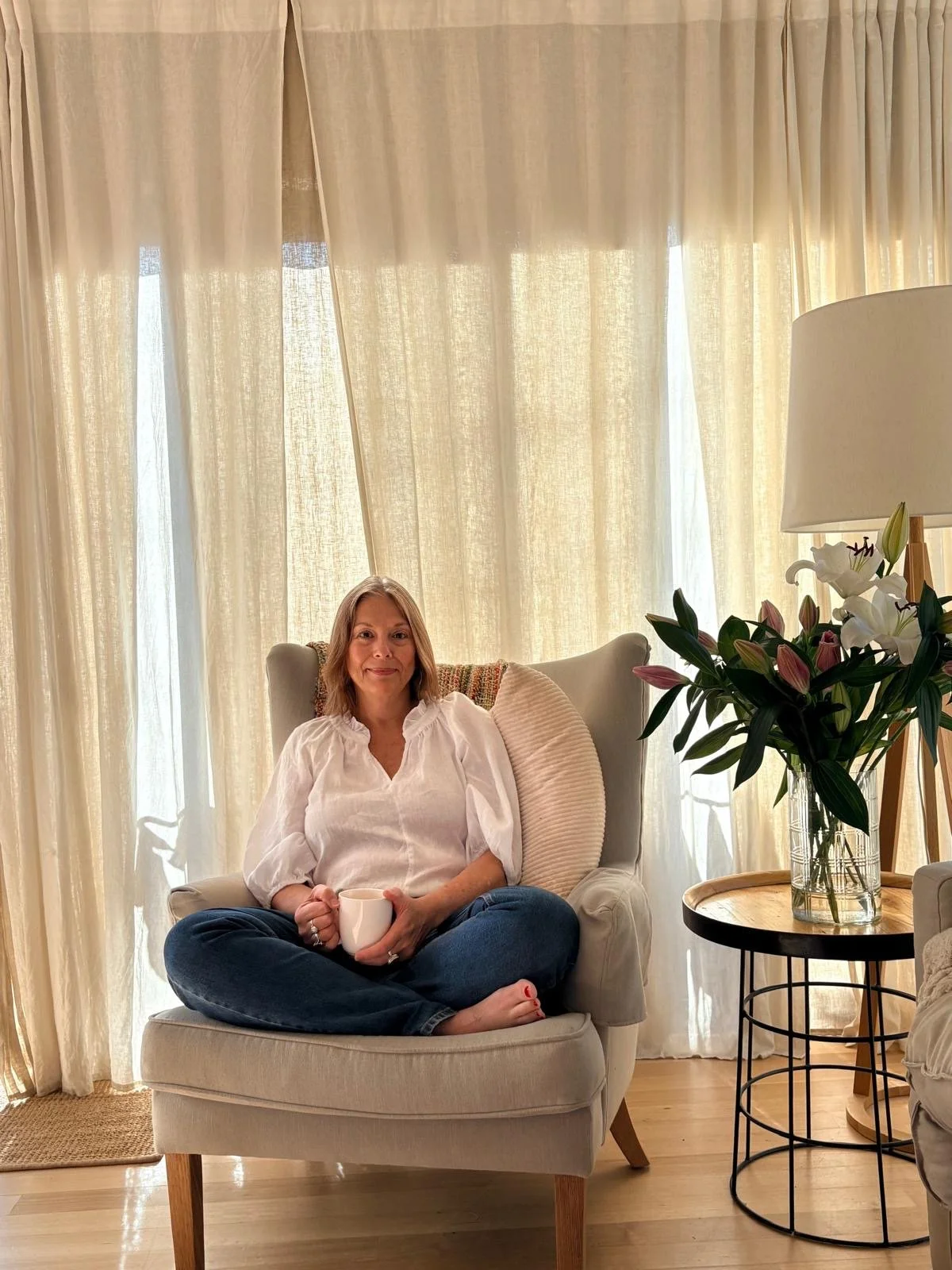 Woman sitting cross-legged on a beige armchair, holding a mug, in a cozy room with cream curtains, a side table with a vase of lilies, and a floor lamp.