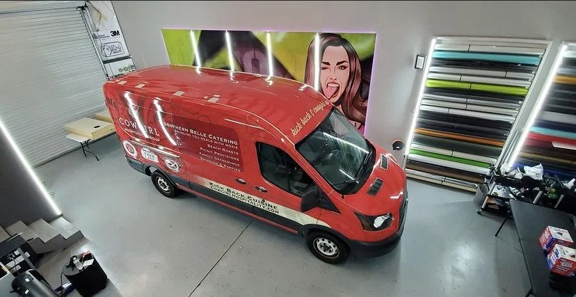 A red catering van parked in an indoor space with wall art and shelves of colored rolls or materials.