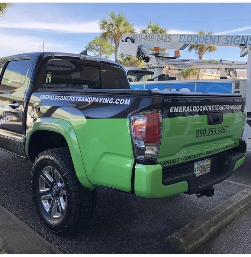 Pickup truck with vibrant green and black graphics advertising concrete and paving services, parked in a lot with a lift and some palm trees in the background.