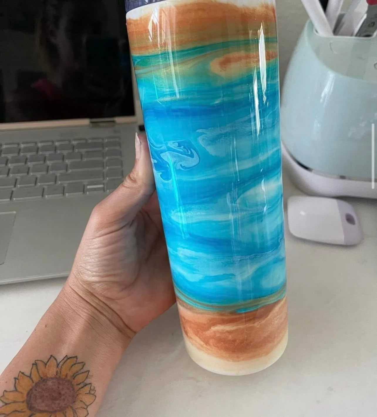 A hand holding a tall, colorful tumbler with a swirling pattern of orange, blue, green, and white, in front of a laptop, a white computer mouse, and a white container holding utensils.