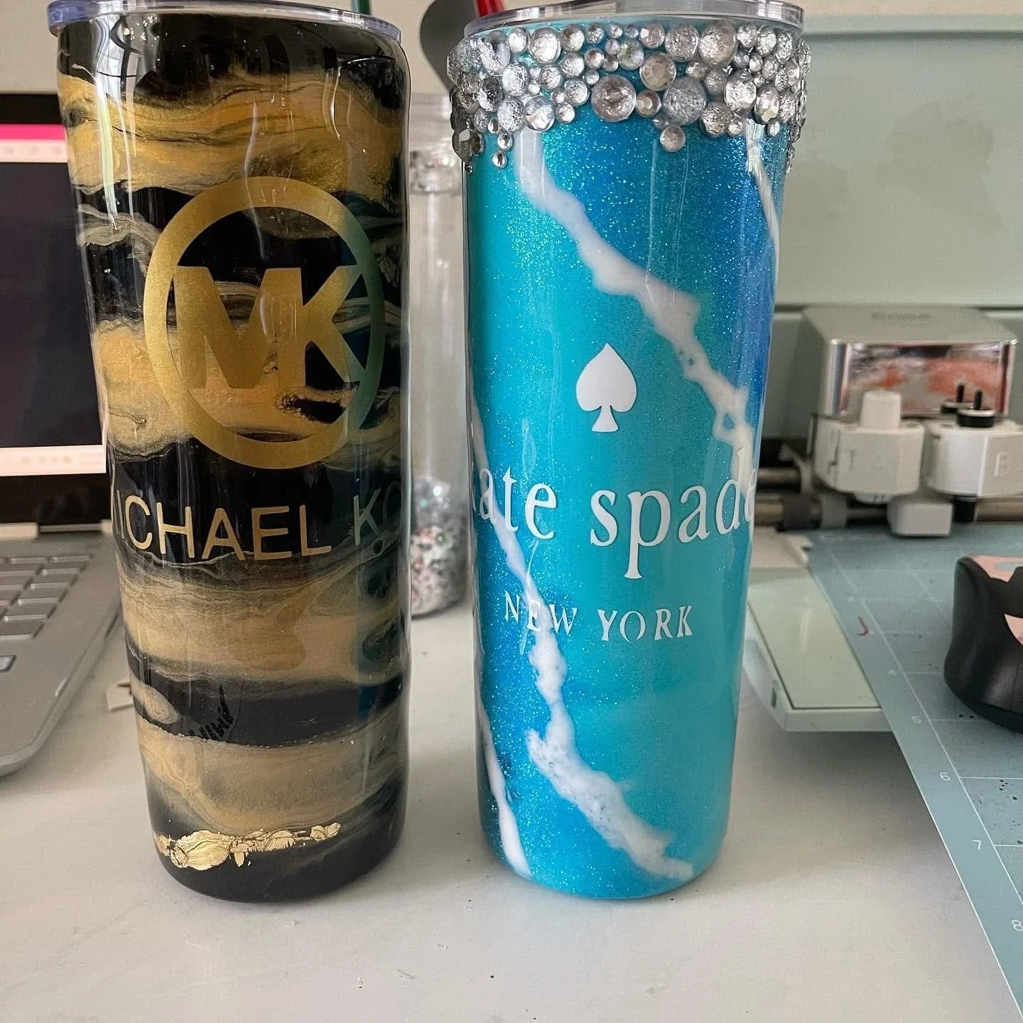 Two large colorful tumblers, one black and gold with the name Michael K. and initial M, the other blue with silver beads on top and white marble pattern, branded with Kate Spade New York.
