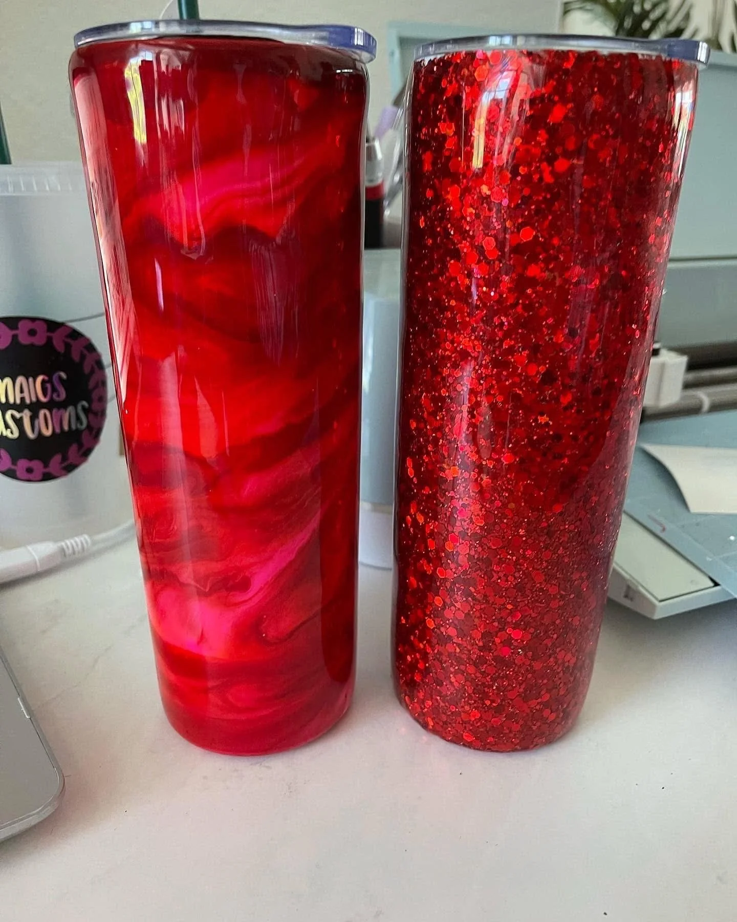 Two tall red decorative glasses, one with a swirled marble pattern and the other covered in red glitter, placed on a white surface.