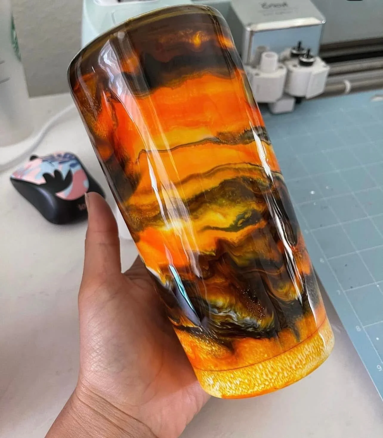A person holding a glass vase with a marbled orange, black, and yellow pattern. In the background, there is a silicone craft mat, a tape measure, and some small bottles.