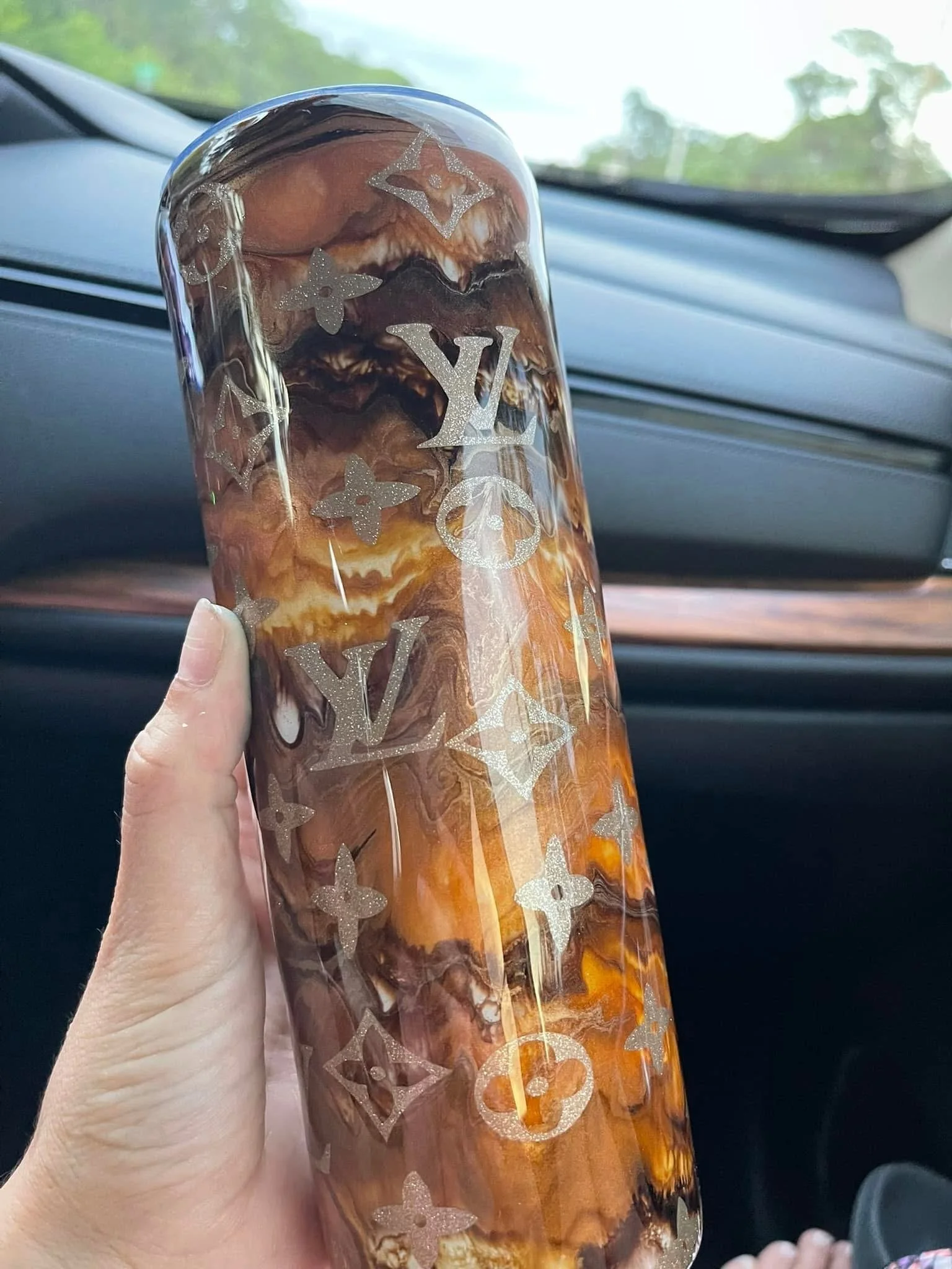 Hand holding a decorated glass with caramel-colored contents inside a car, with trees visible through the windshield.