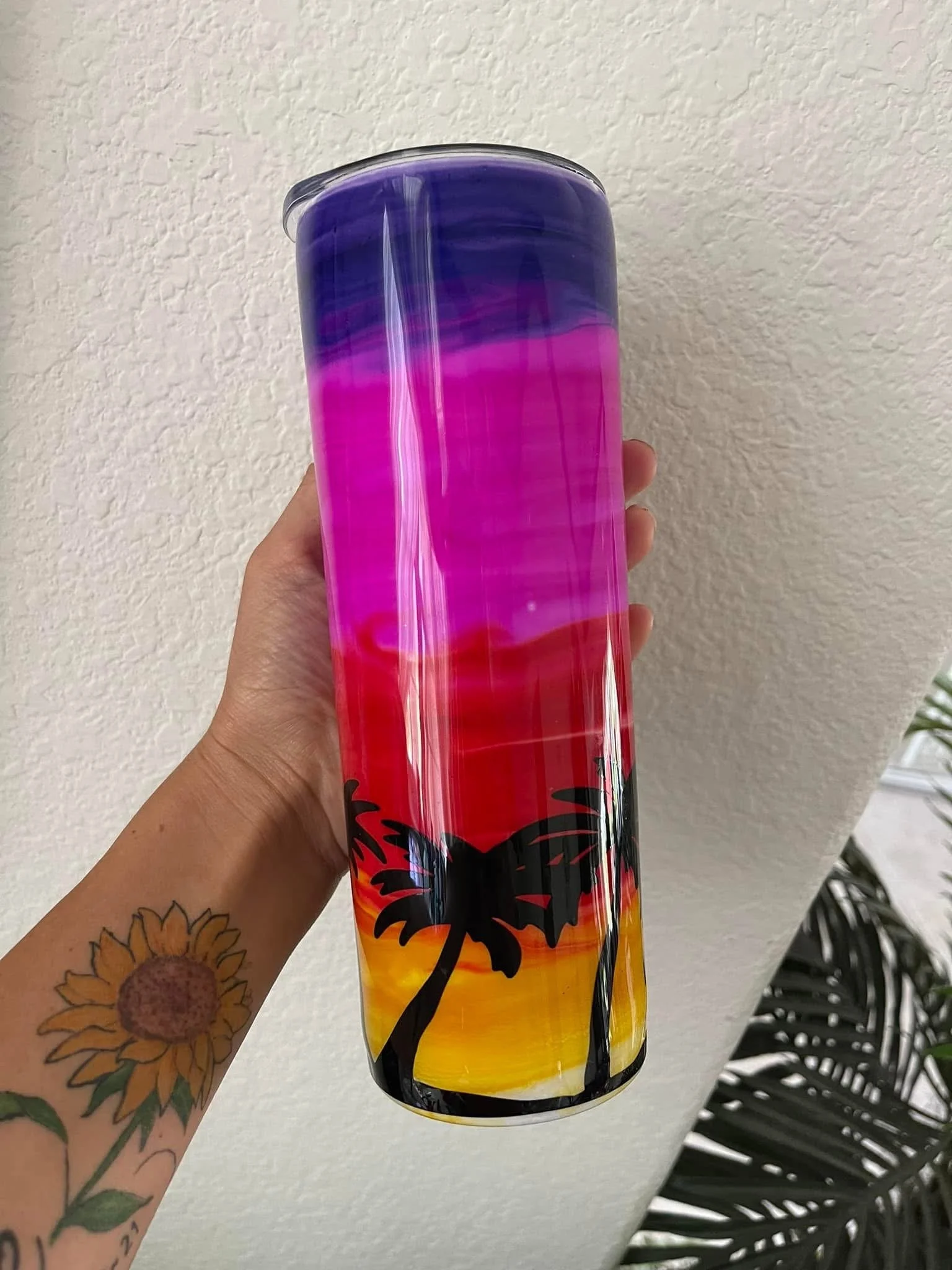 Hand holding a colorful, tie-dye style tumbler with a palm tree design against a textured white wall background.