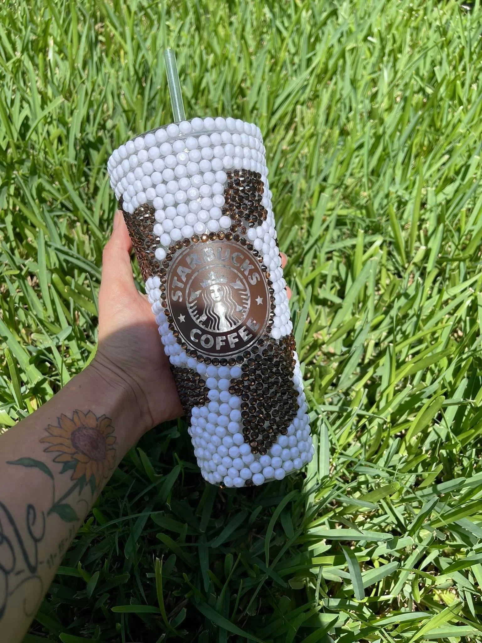 Person holding a Starbucks coffee cup decorated with white and black beads, with a straw in a grassy outdoor setting.