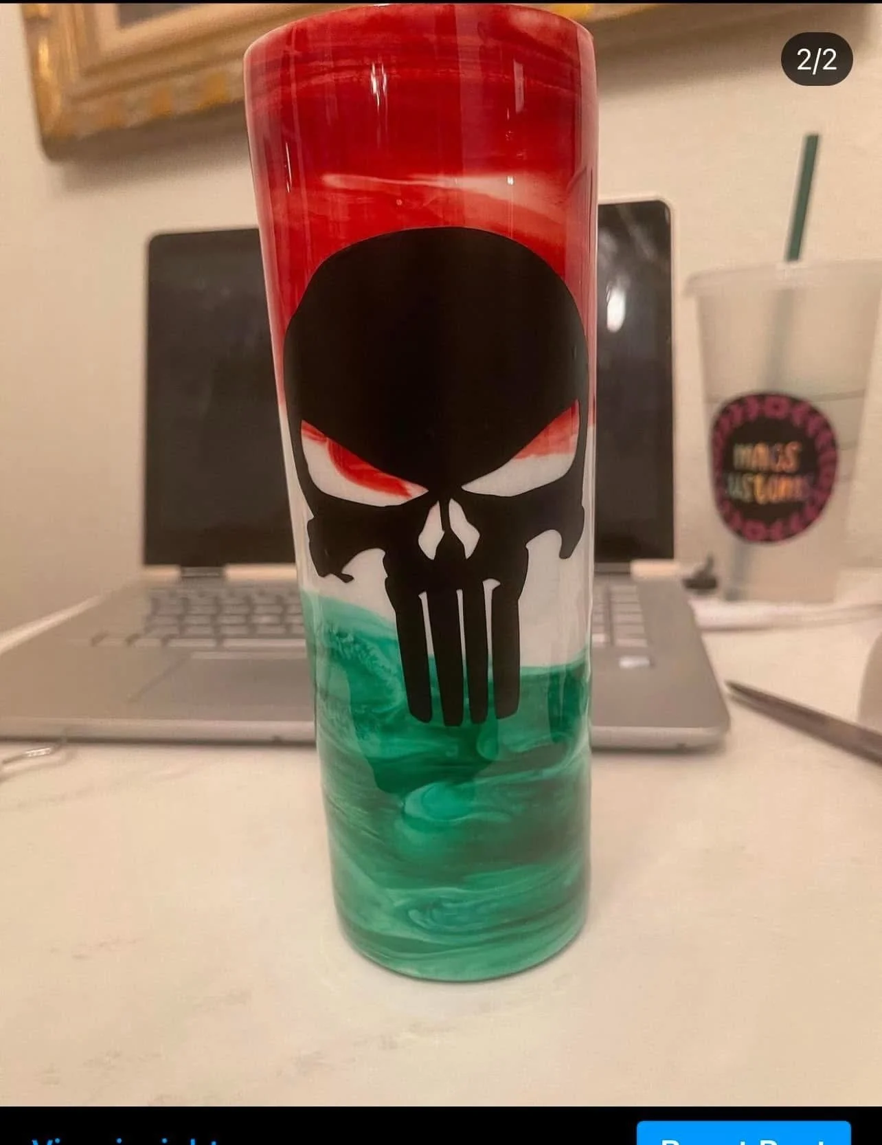 A tall, colorful tumbler with a black skull design and green, white, red, and black swirls, placed on a desk with a laptop, a drink cup, and a pen in the background.