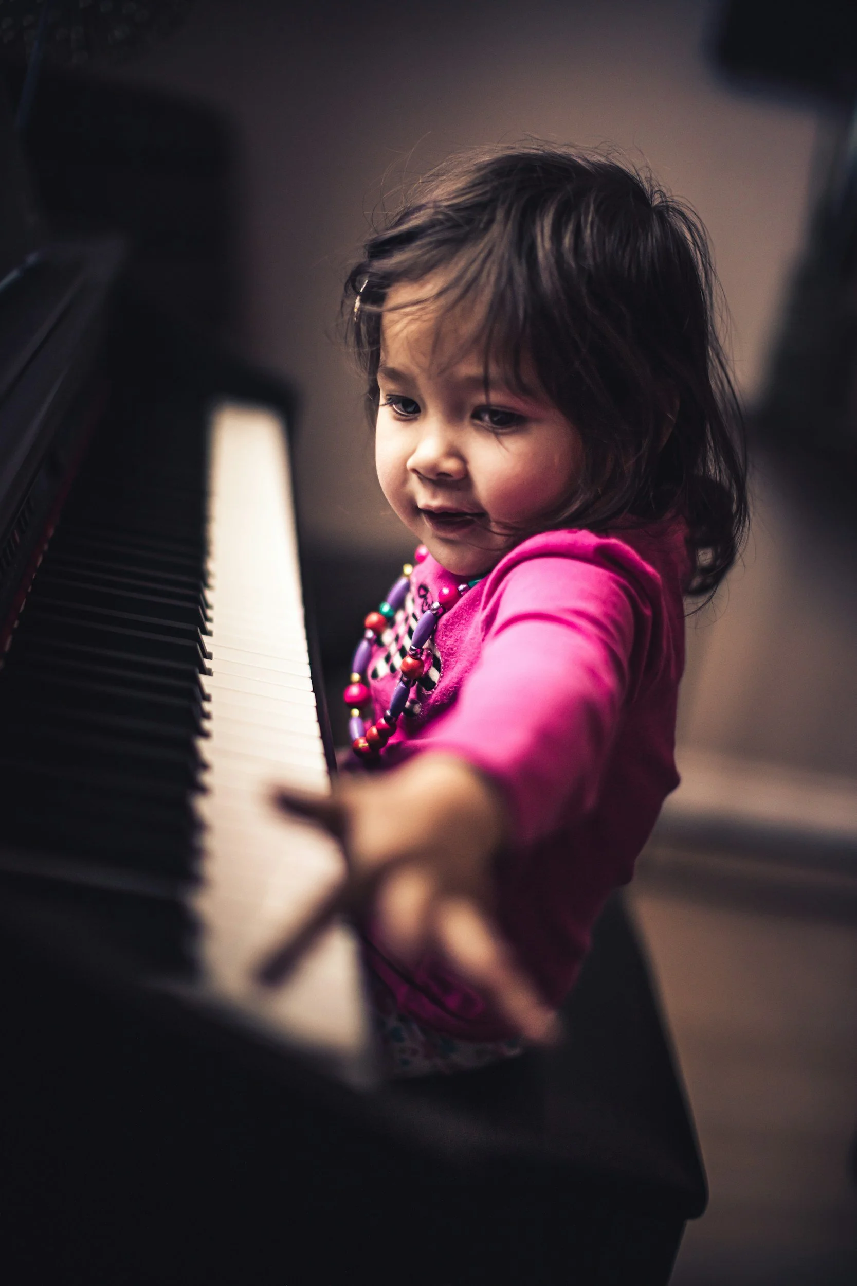 Young girl with dark hair wearing a pink shirt and colorful bead necklace playing piano, focus on her face and hands.