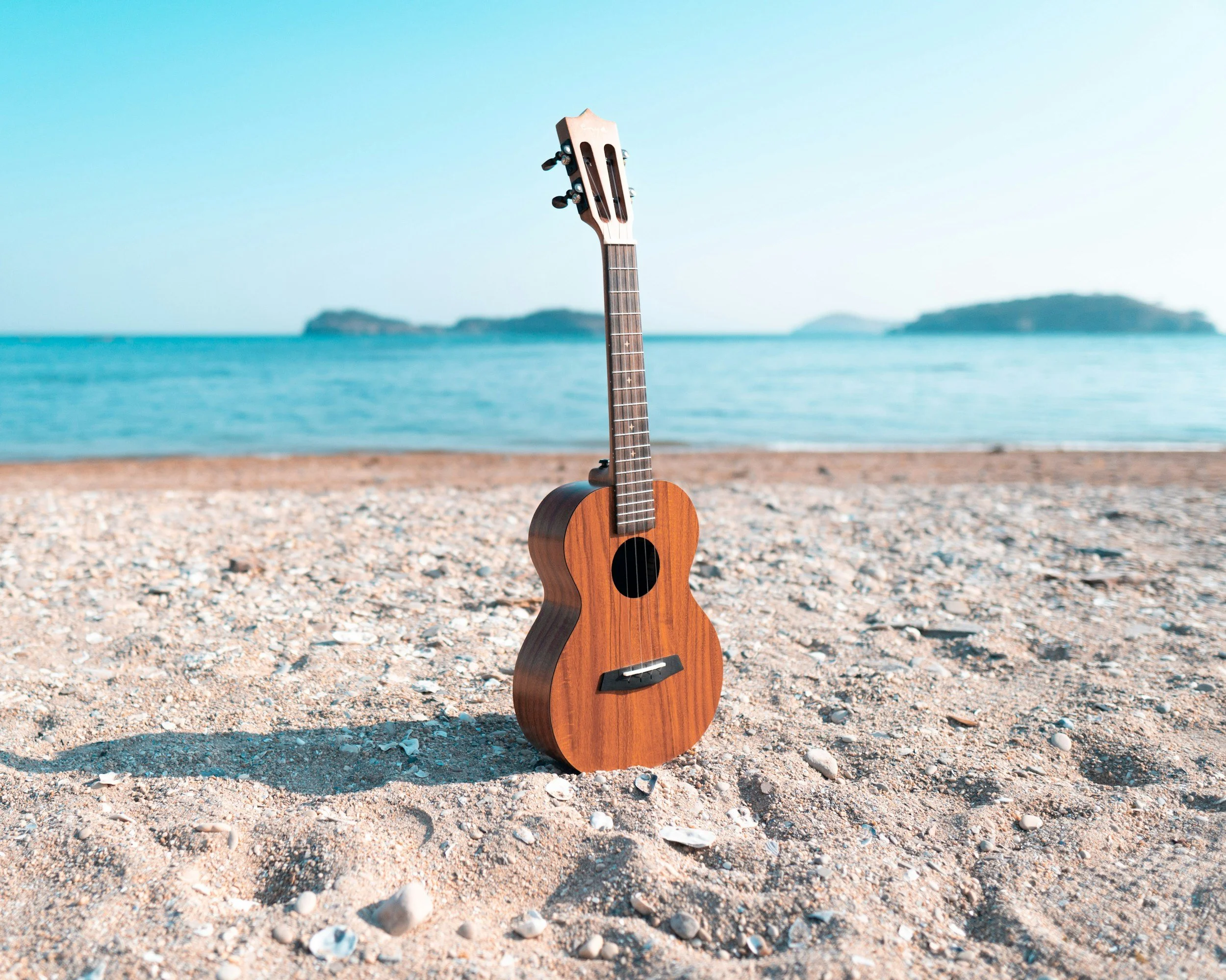 A small wooden ukulele standing upright on a sandy beach near the water with small islands in the background.