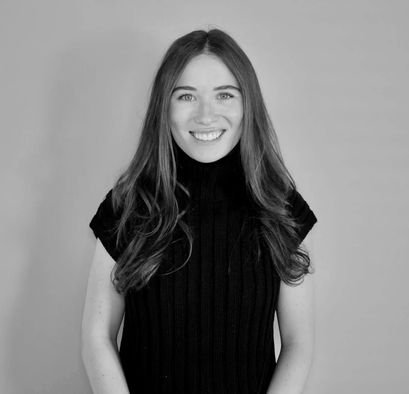 Black and white portrait of a woman with long, wavy hair, wearing a sleeveless, ribbed, turtleneck top, smiling, standing against a plain background.