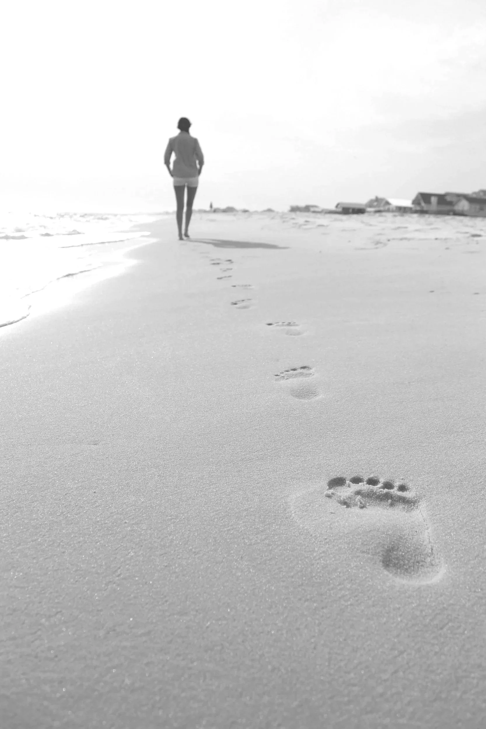 A person walking alone on a sandy beach, leaving a trail of footprints behind them, with buildings in the distance and a partly cloudy sky.