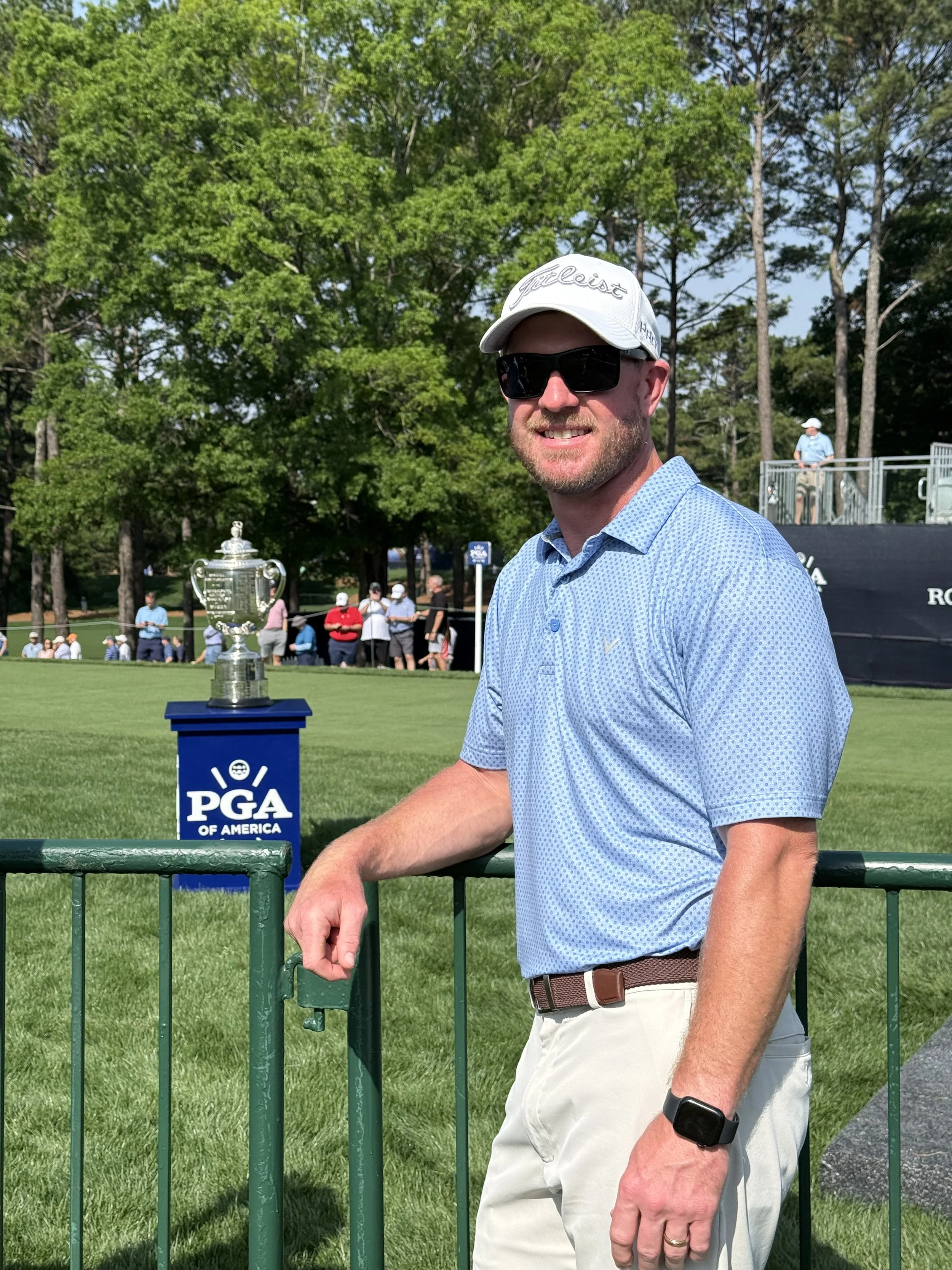 A man in a golf shirt, white pants, wearing sunglasses and a white golf cap, standing outdoors next to a green golf course fence with a trophy and a PGA of America sign behind him.