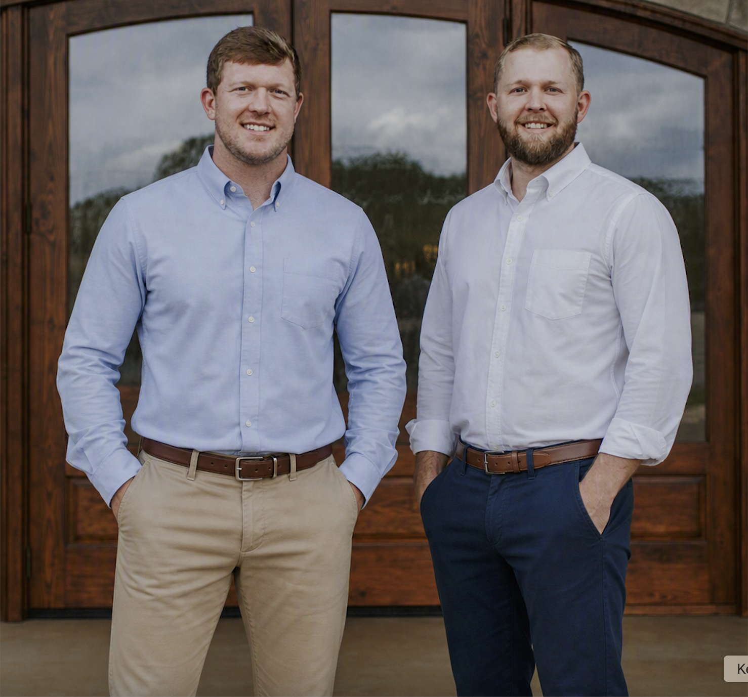 Two smiling men standing outside in front of a wooden doorway, wearing button-up shirts and khakis or trousers, with hands in pockets.