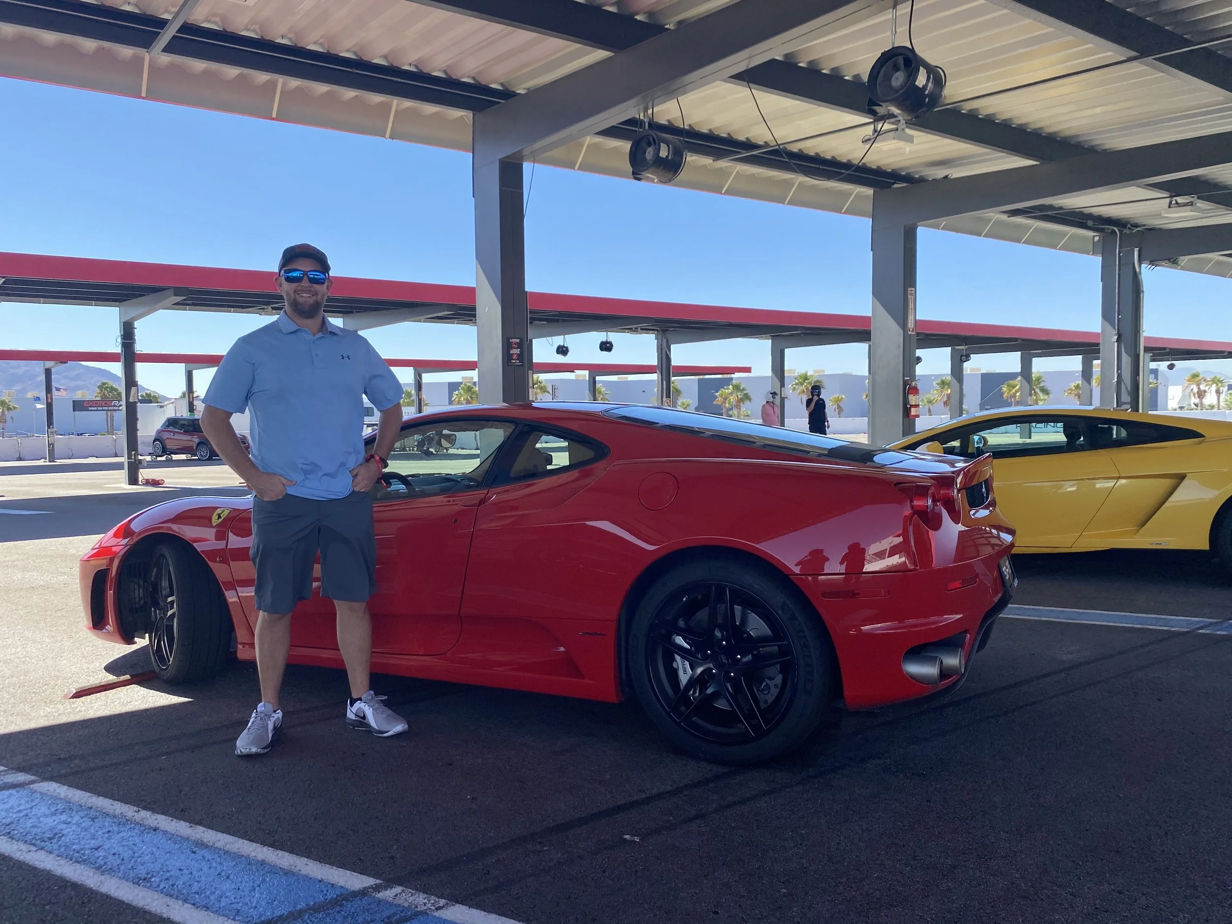 A man wearing a blue shirt, shorts, sunglasses, and a cap stands next to a red sports car at a parking lot with a canopy overhead. There are yellow cars parked nearby and a few people in the background, along with palm trees and buildings under a clear blue sky.