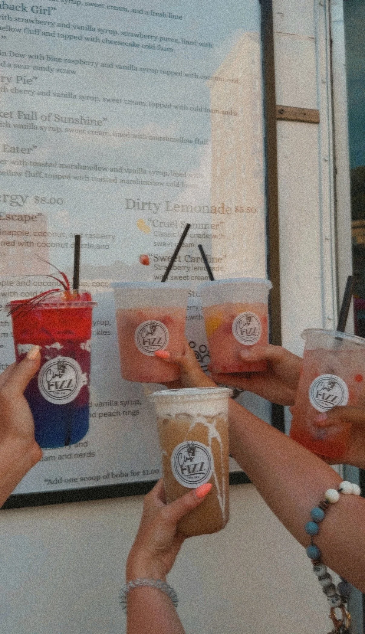 Six hands holding various colorful drinks with straws, in front of a menu board that has dessert options. The drinks are in plastic cups with a logo, and the hands are visible with bracelets and nail polish.