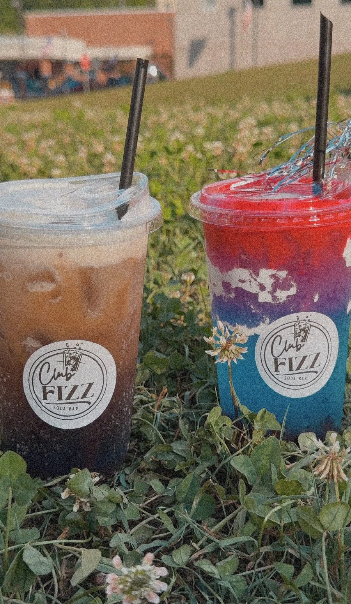 Two colorful soda cups with black straws from Club Fizz resting on grass, with a blurred background of a building and people.