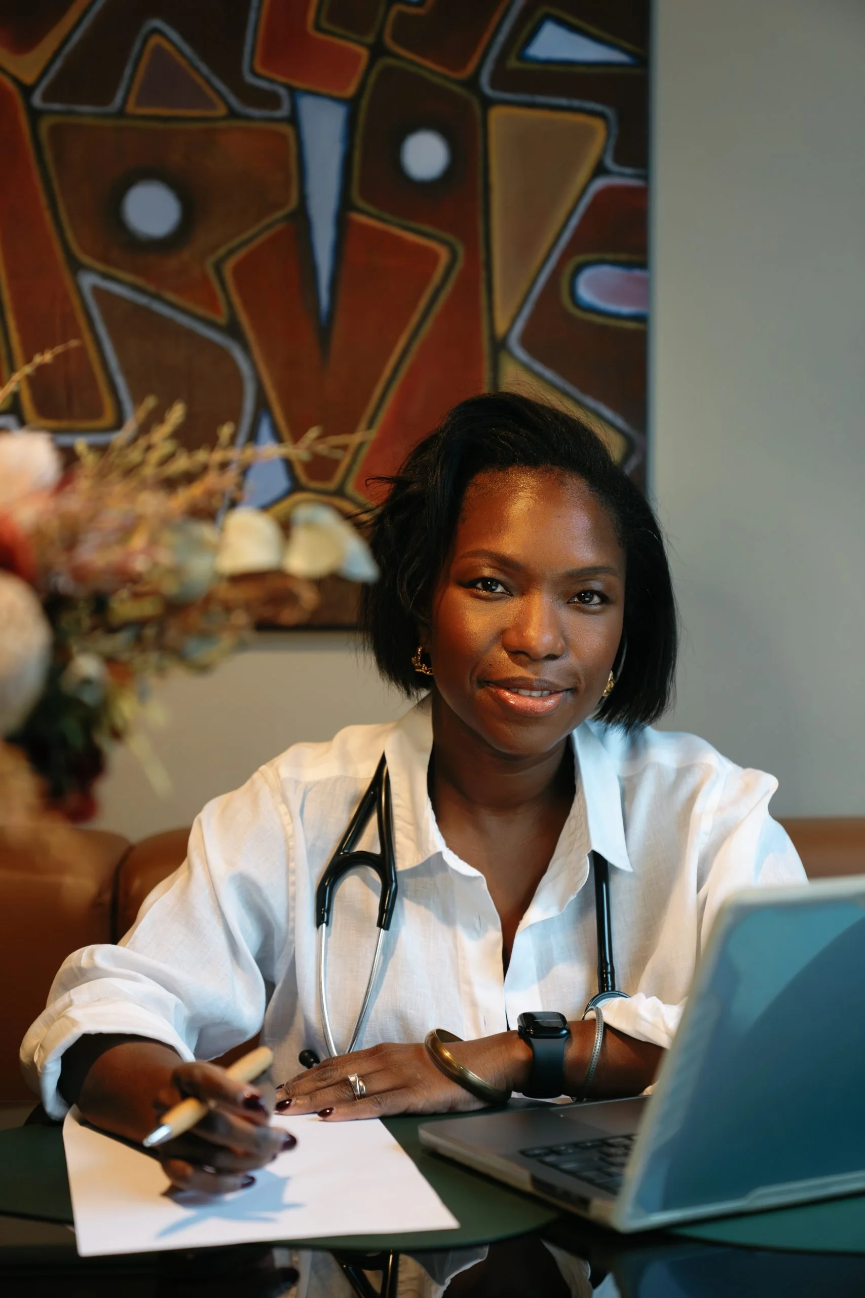A woman dressed as a healthcare professional, possibly a doctor, sitting at a desk with a laptop, phone, and a stethoscope around her neck, smiling and looking at the camera. Behind her is a colorful geometric artwork.