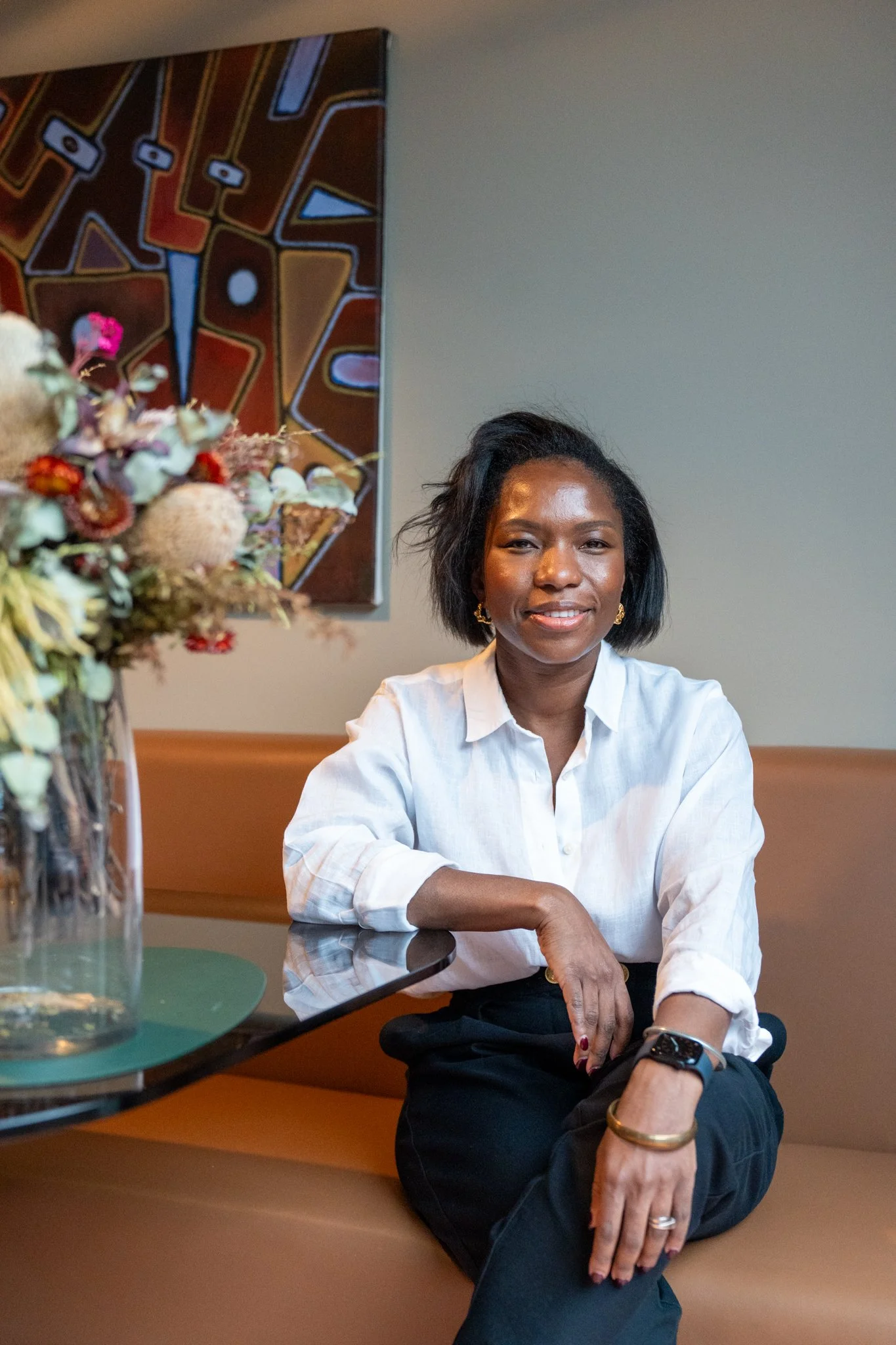 A woman with dark hair, wearing a white shirt and accessories, sitting at a table with a floral arrangement in a glass vase in a room with a colorful abstract painting on the wall.