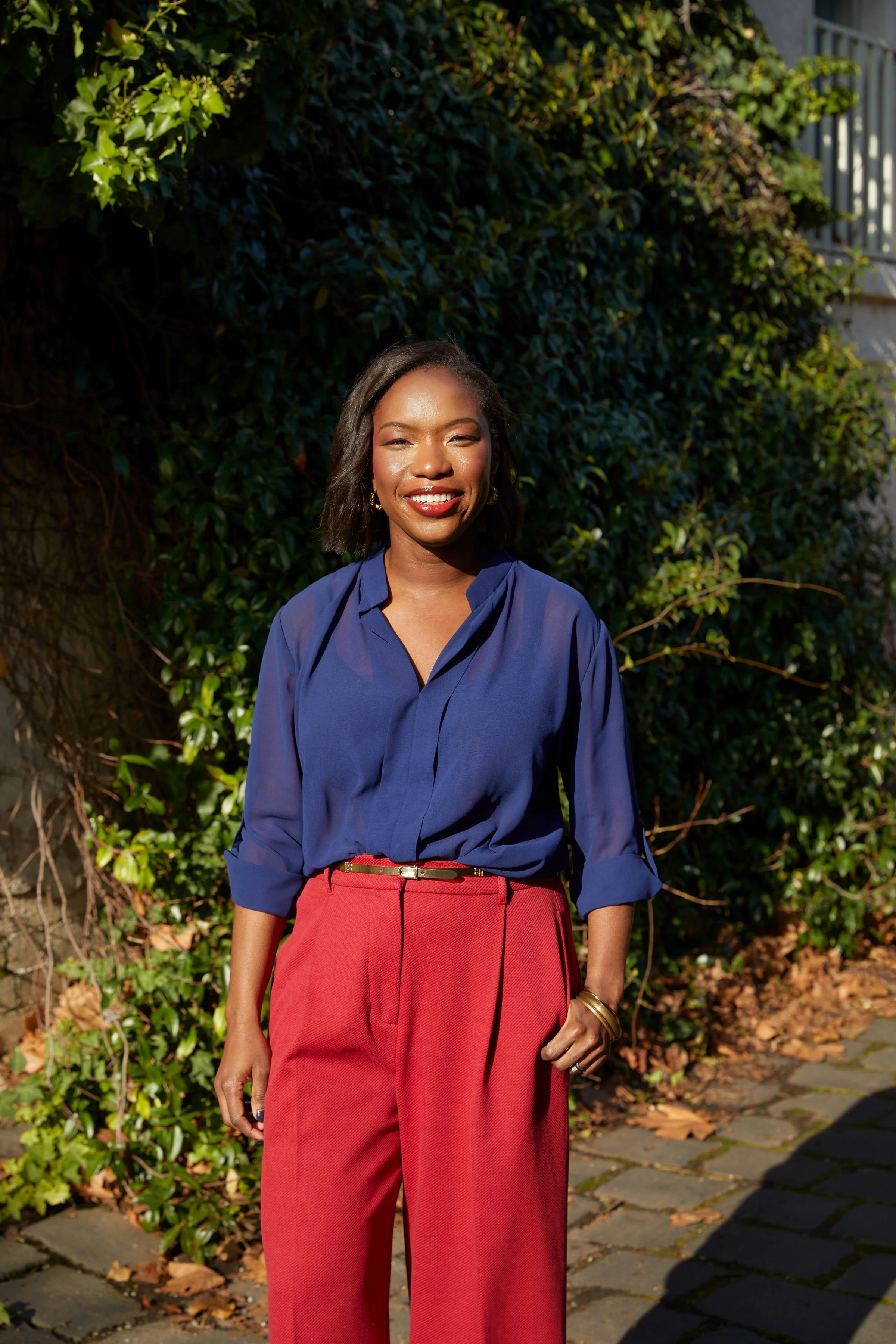 A woman standing outdoors on a sunny day, wearing a blue blouse and red trousers, with a background of green foliage and fallen leaves.