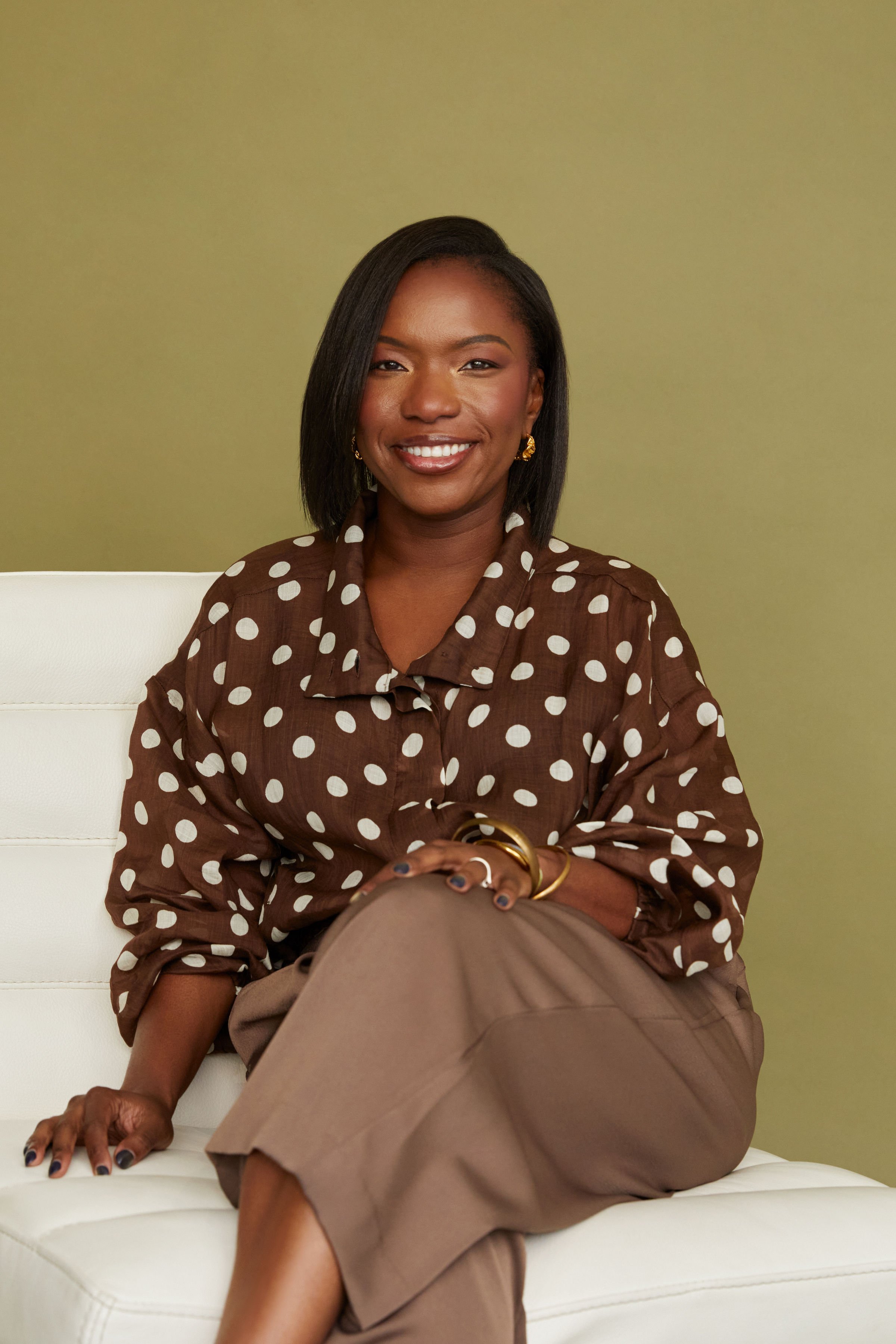 A woman sitting on a white cushioned bench, smiling, wearing a brown polka dot blouse and beige pants, with a greenish wall behind her.