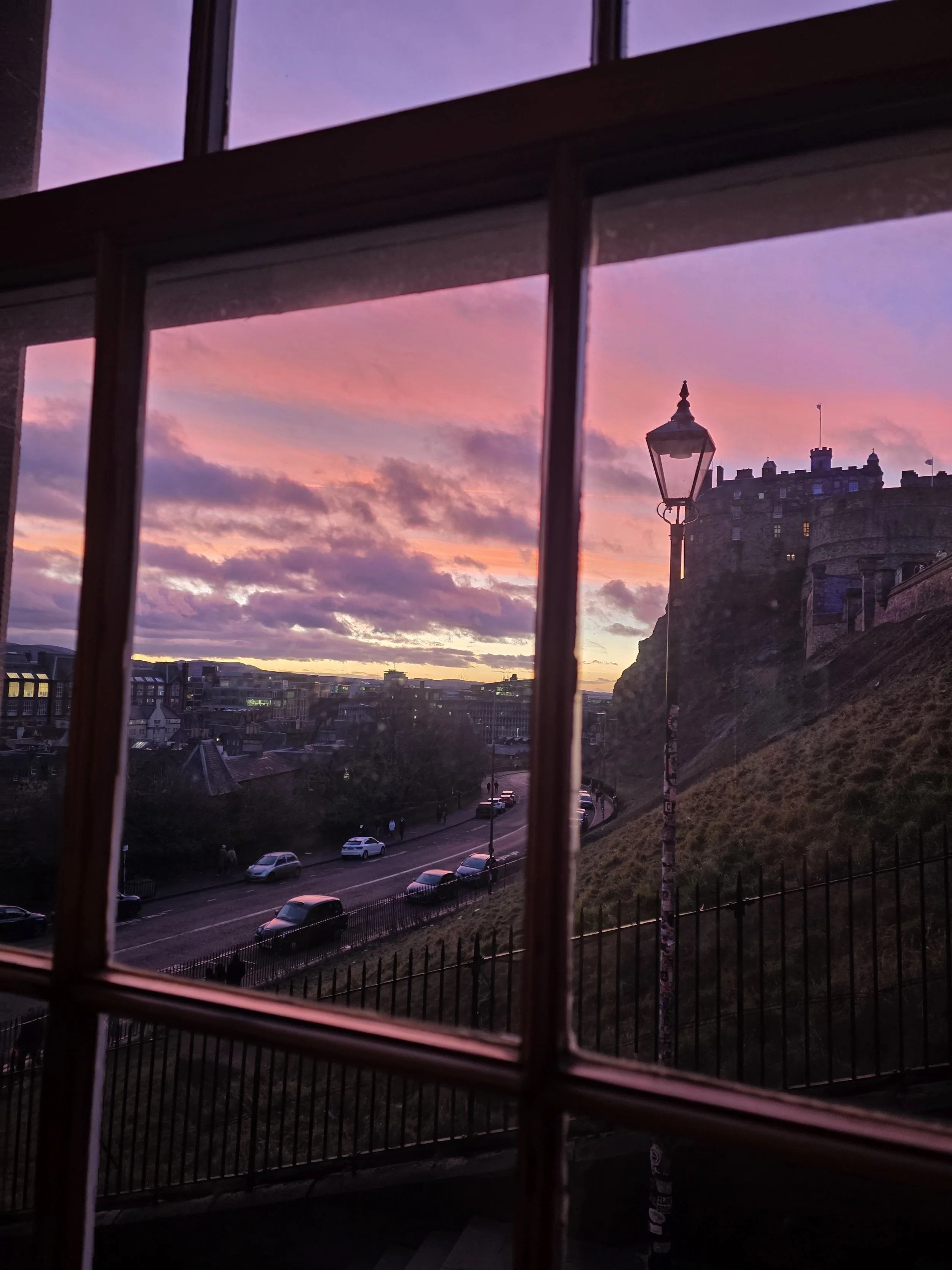 View of a cityscape during sunset seen through window with grid bars, with a historic castle and a street with parked cars in the foreground.