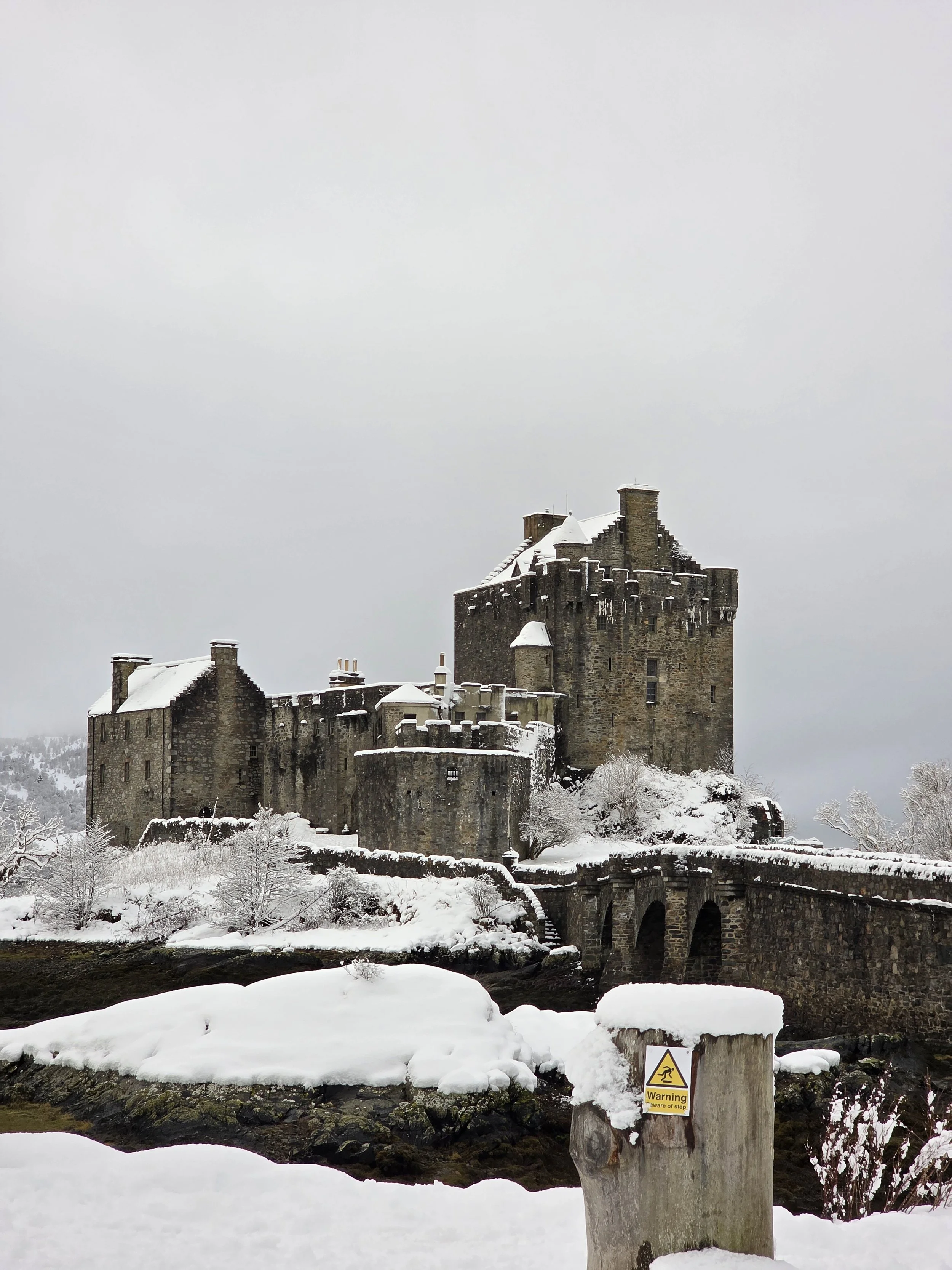 Snow-covered ancient stone castle on a hill with a moat and warning sign in the foreground, under a cloudy sky.