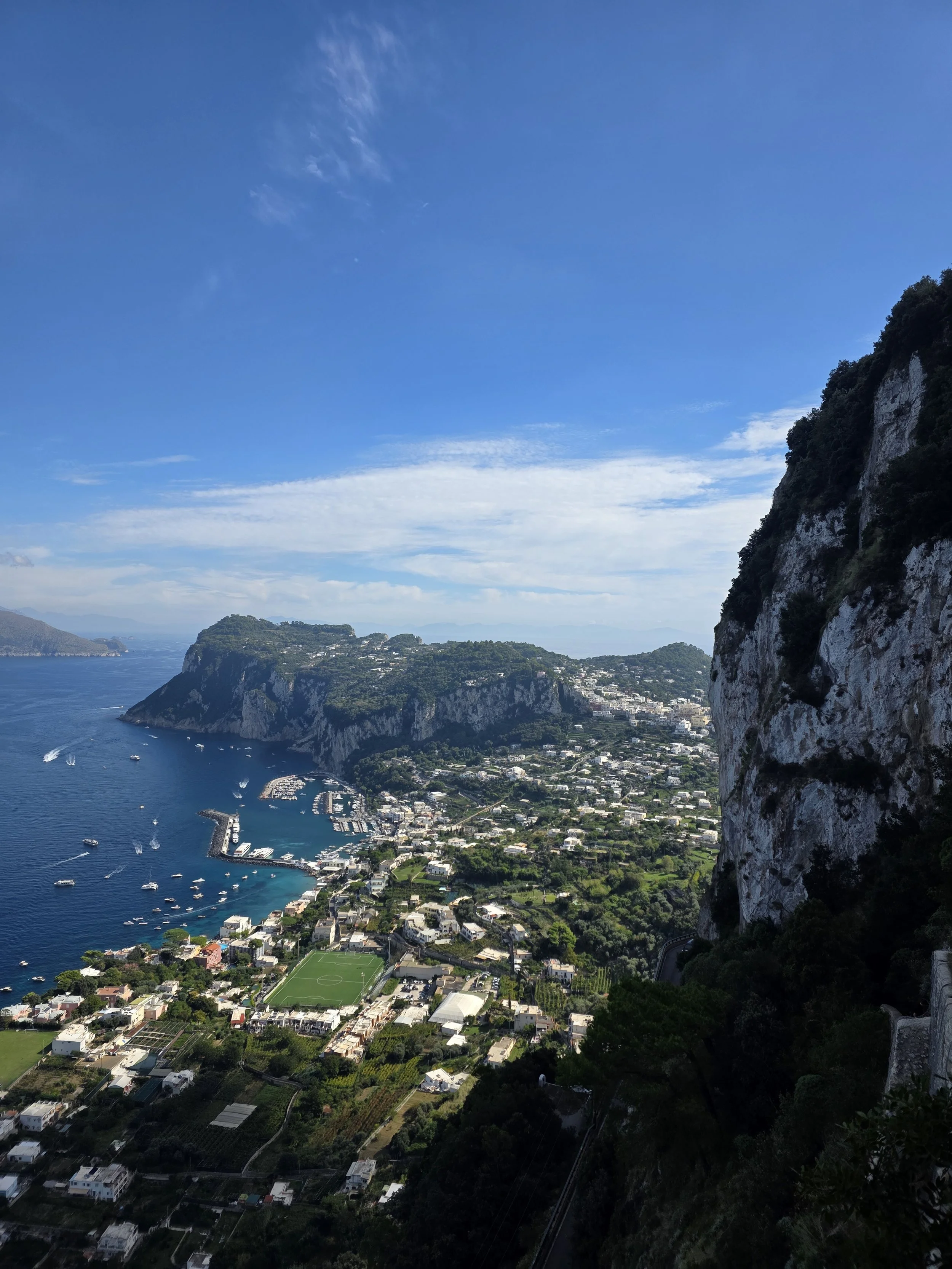 Scenic view of a coastal town with a harbor, boats, and a sports field, surrounded by lush green hills and cliffs under a bright blue sky.