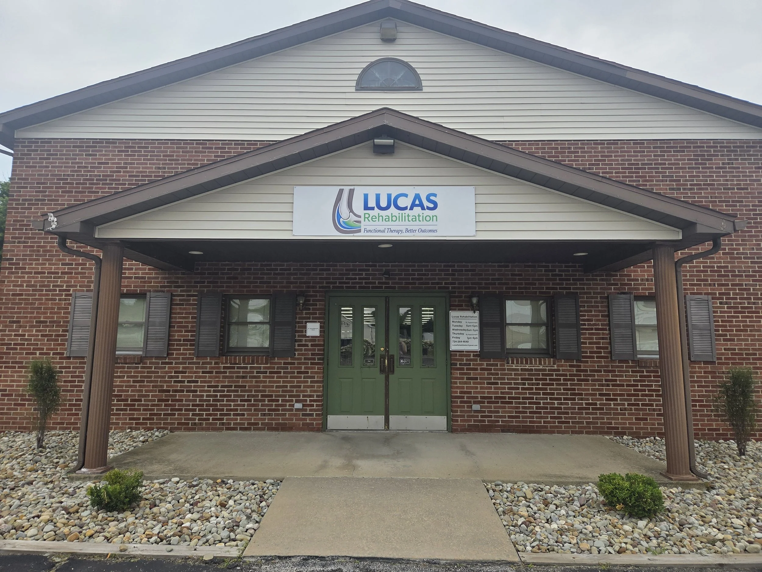 Front entrance of Lucas Rehabilitation building with brick walls and beige siding, green door, and a sign that reads "Lucas Rehabilitation" with services and hours listed.