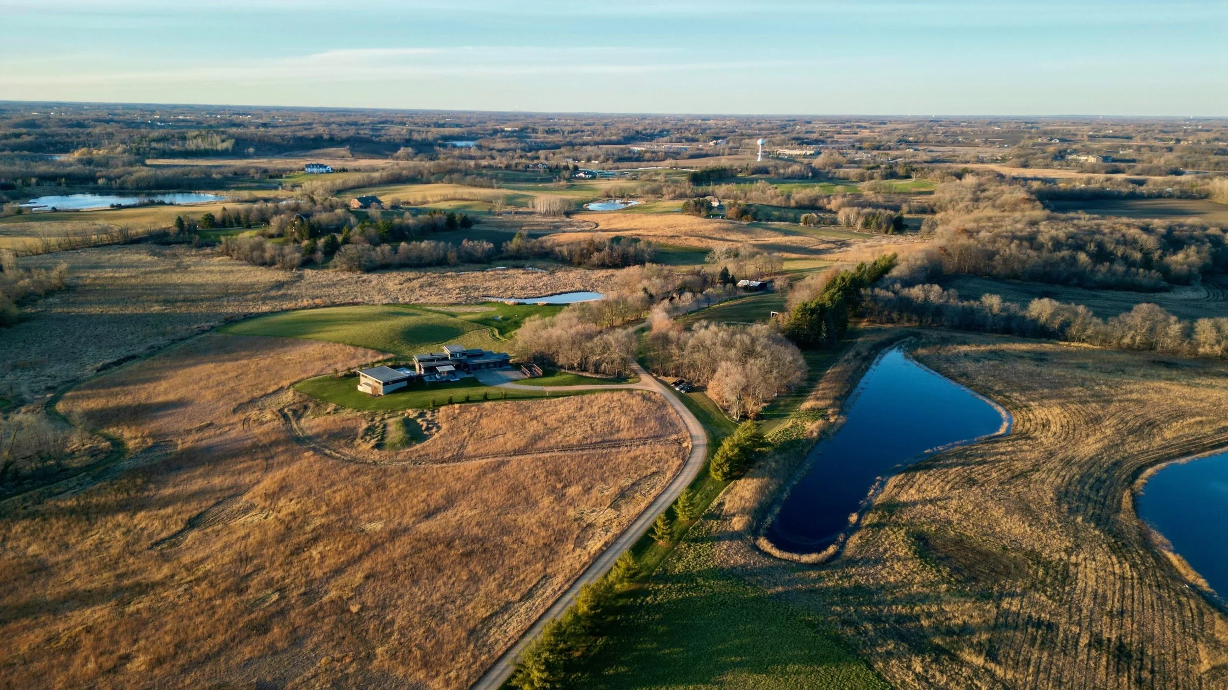 Aerial view of a rural landscape with open fields, ponds, a golf course, and a few buildings, under a clear sky.