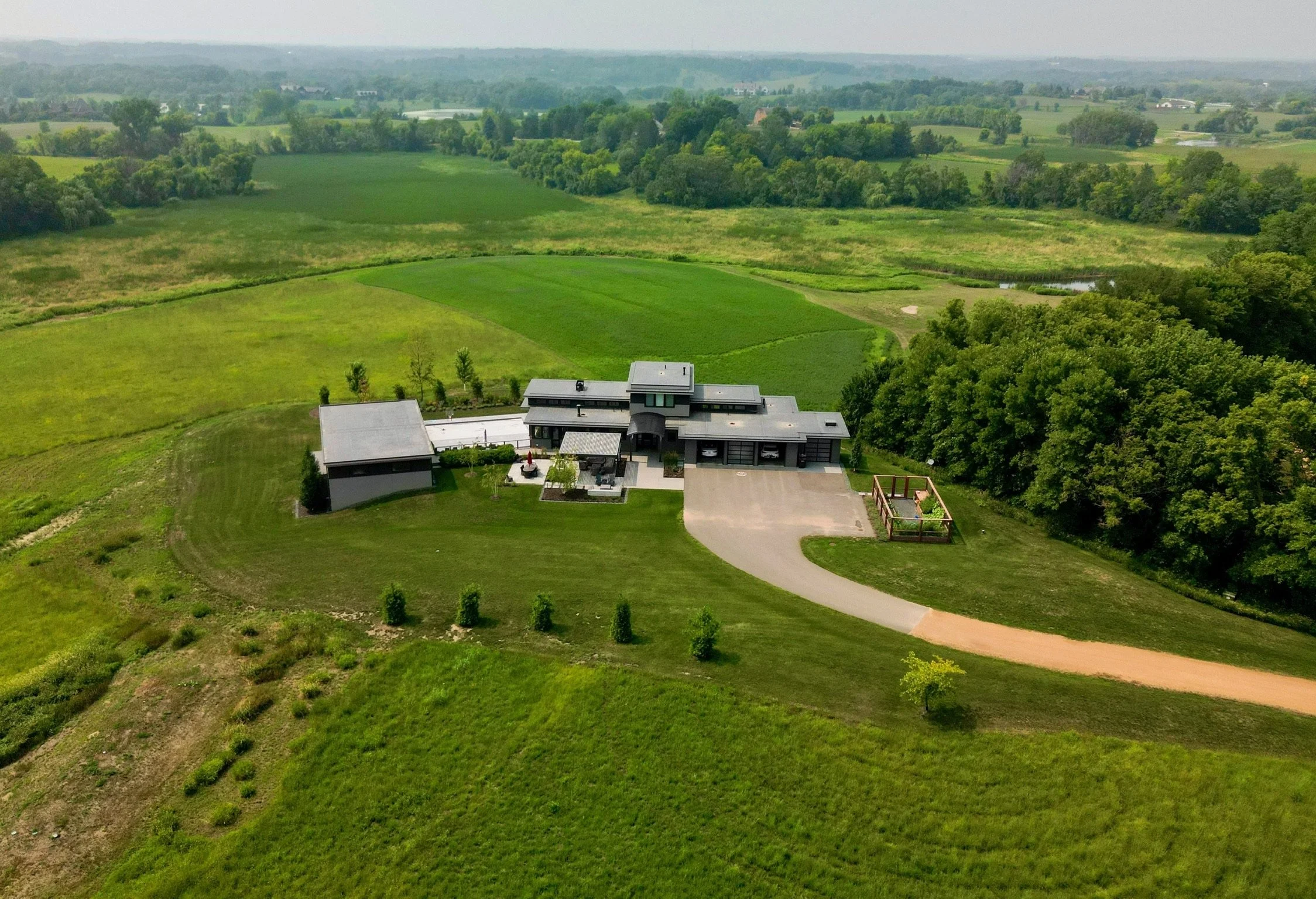 Aerial view of a modern house with a large driveway and surrounding lush green landscape, including fields and trees, with hills in the background.