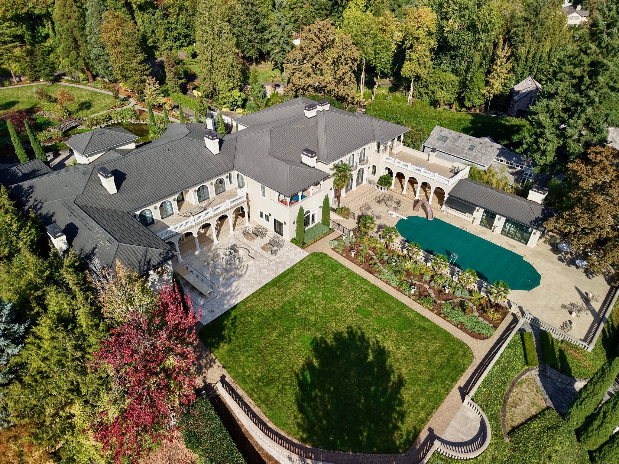Aerial view of a luxurious estate with a large house, swimming pool, lush green lawn, and surrounding tall trees.
