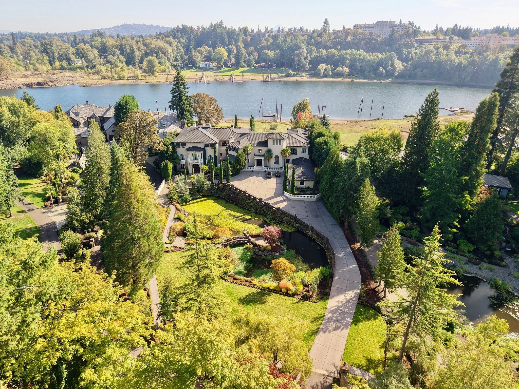 Aerial view of a large house with a driveway, surrounded by tall trees, near a body of water with sailboats and a forested hillside in the background.