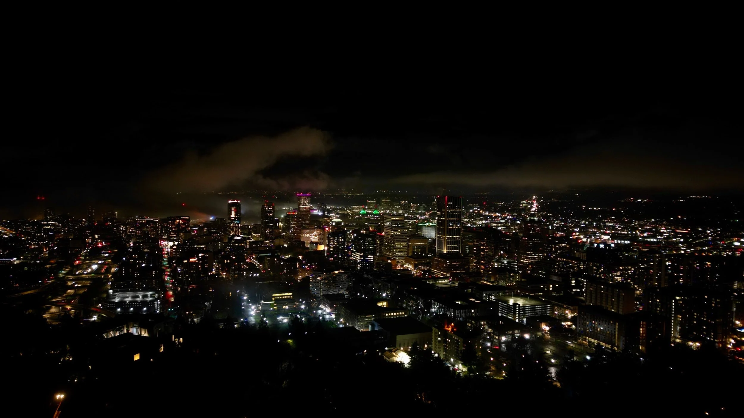 Nighttime city skyline with illuminated buildings and smoke or fog in the background.