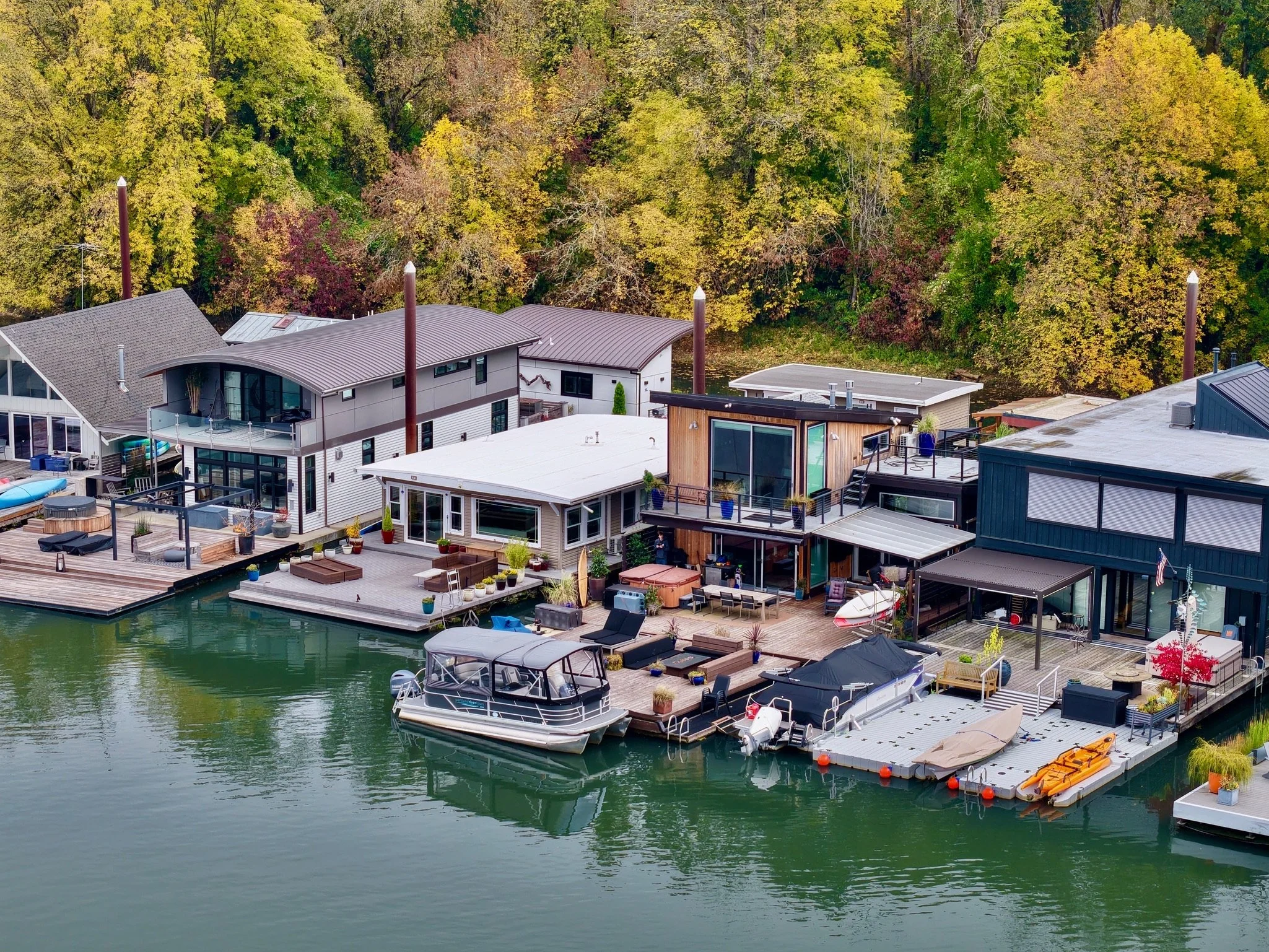 Aerial view of houses with decks by a body of water; boats anchored at docks; trees with green and fall foliage in the background.