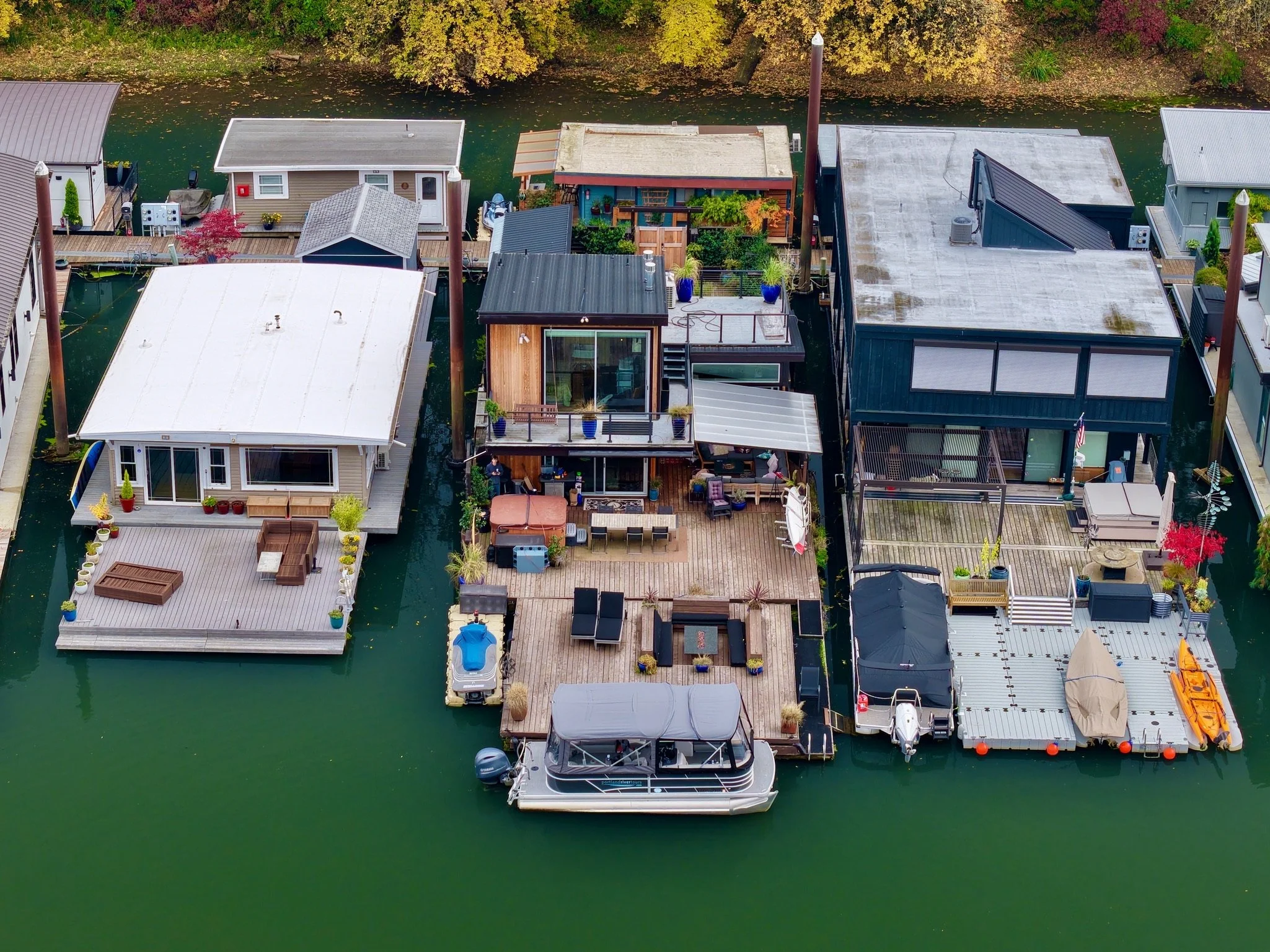 An aerial view of a neighborhood with houses built on water, connected by wooden decks. A boat is docked at a house in the foreground, and there are several colorful potted plants and outdoor furniture on the decks. Trees with autumn foliage line the