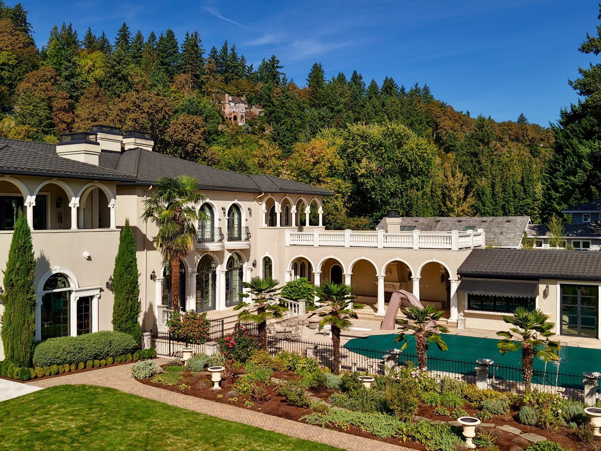 Luxury white house with arched windows, surrounded by lush greenery and trees, set against a backdrop of a densely wooded hillside with large houses visible among the trees under a bright blue sky.