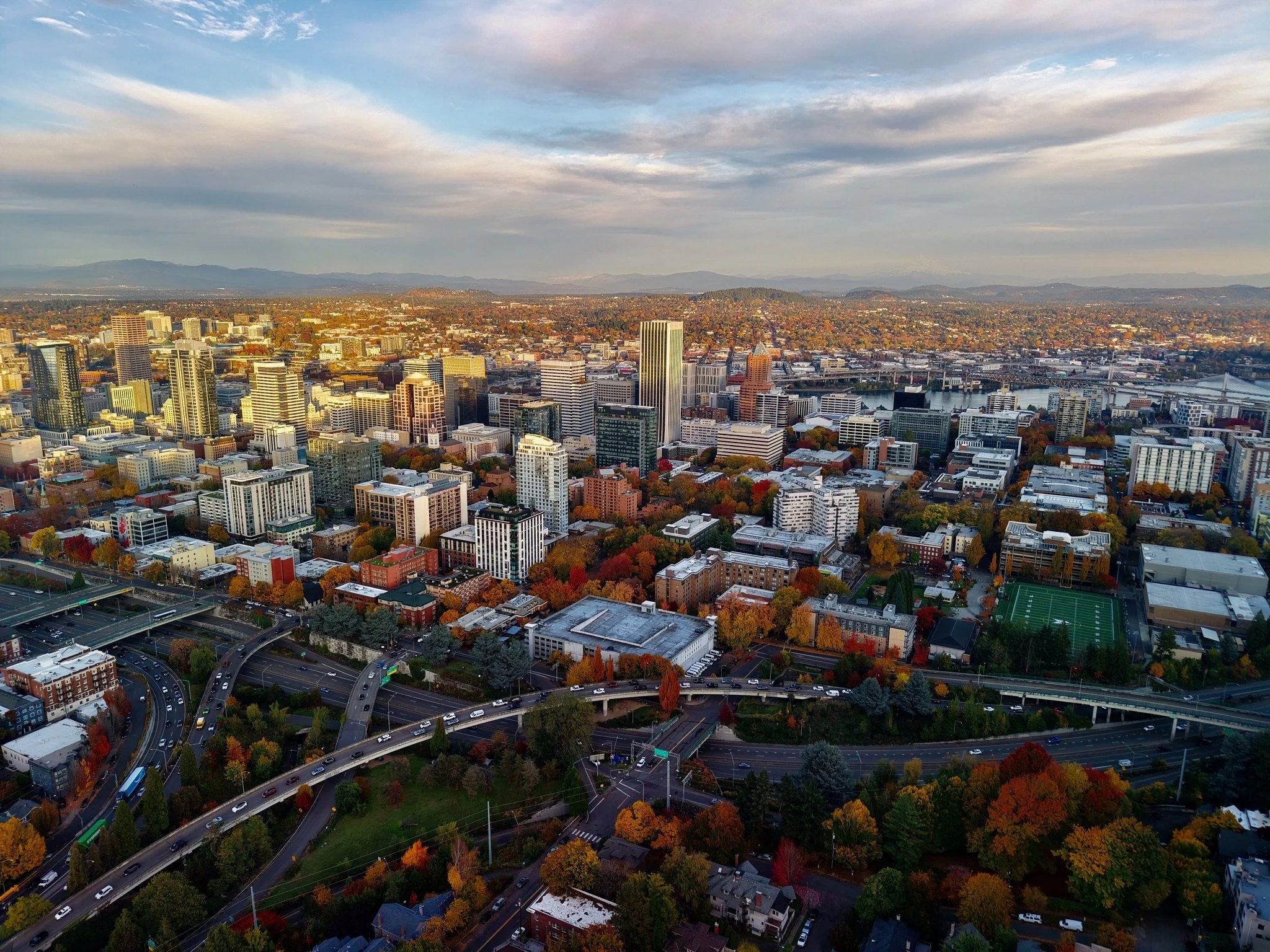 Aerial view of a city skyline with tall buildings, a river, and highways, with mountains in the background and trees with autumn foliage in the foreground.