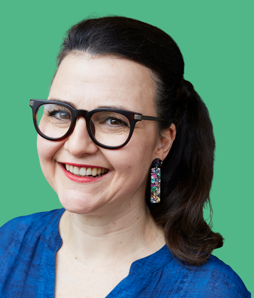 A smiling woman with dark hair tied back, wearing large black glasses, colorful earrings, and a blue top, against a green background.