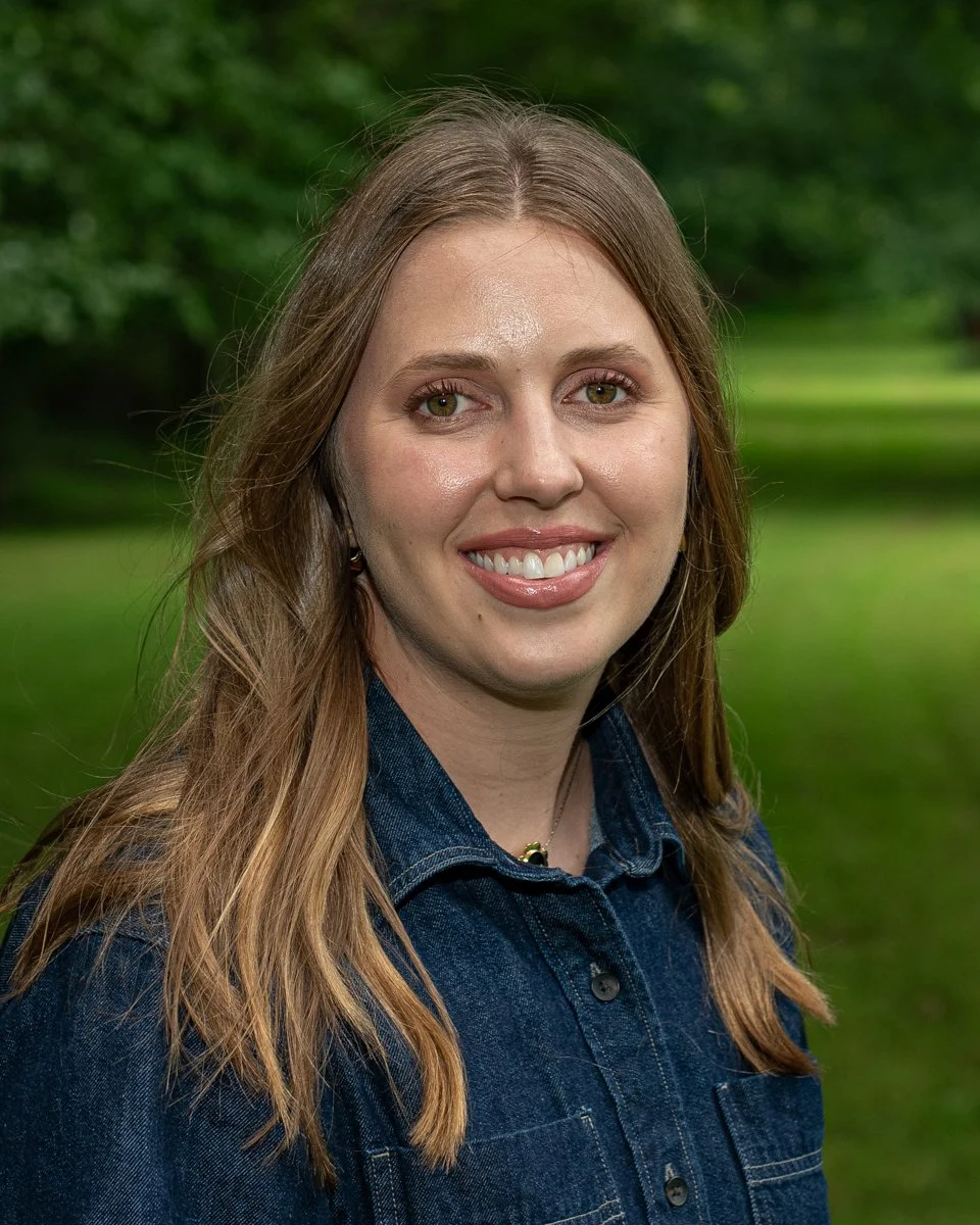 A woman with long light brown hair and light skin, smiling, wearing a denim jacket, outdoors with green trees in the background.