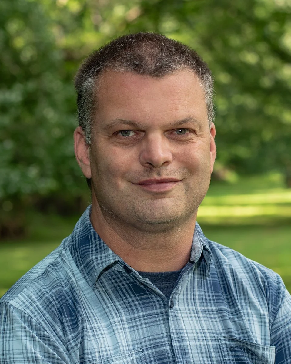 Portrait of a man with short dark hair and light skin, wearing a blue plaid shirt, standing outdoors with blurred green trees in the background.