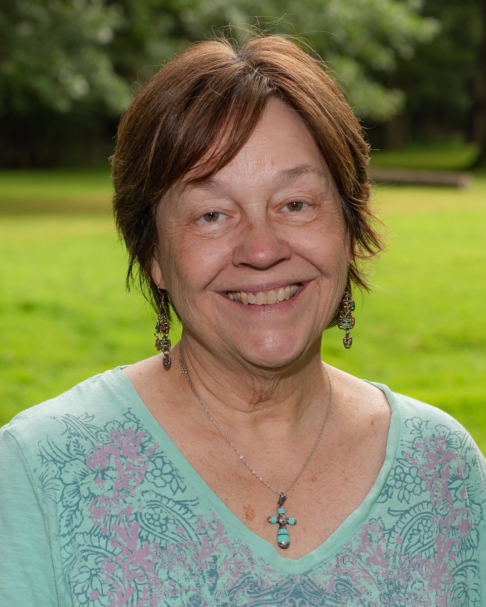 A smiling older woman with short brown hair standing outdoors in a grassy park with trees in the background. She is wearing a light blue top with floral patterns, earrings, and a necklace with a turquoise cross pendant.