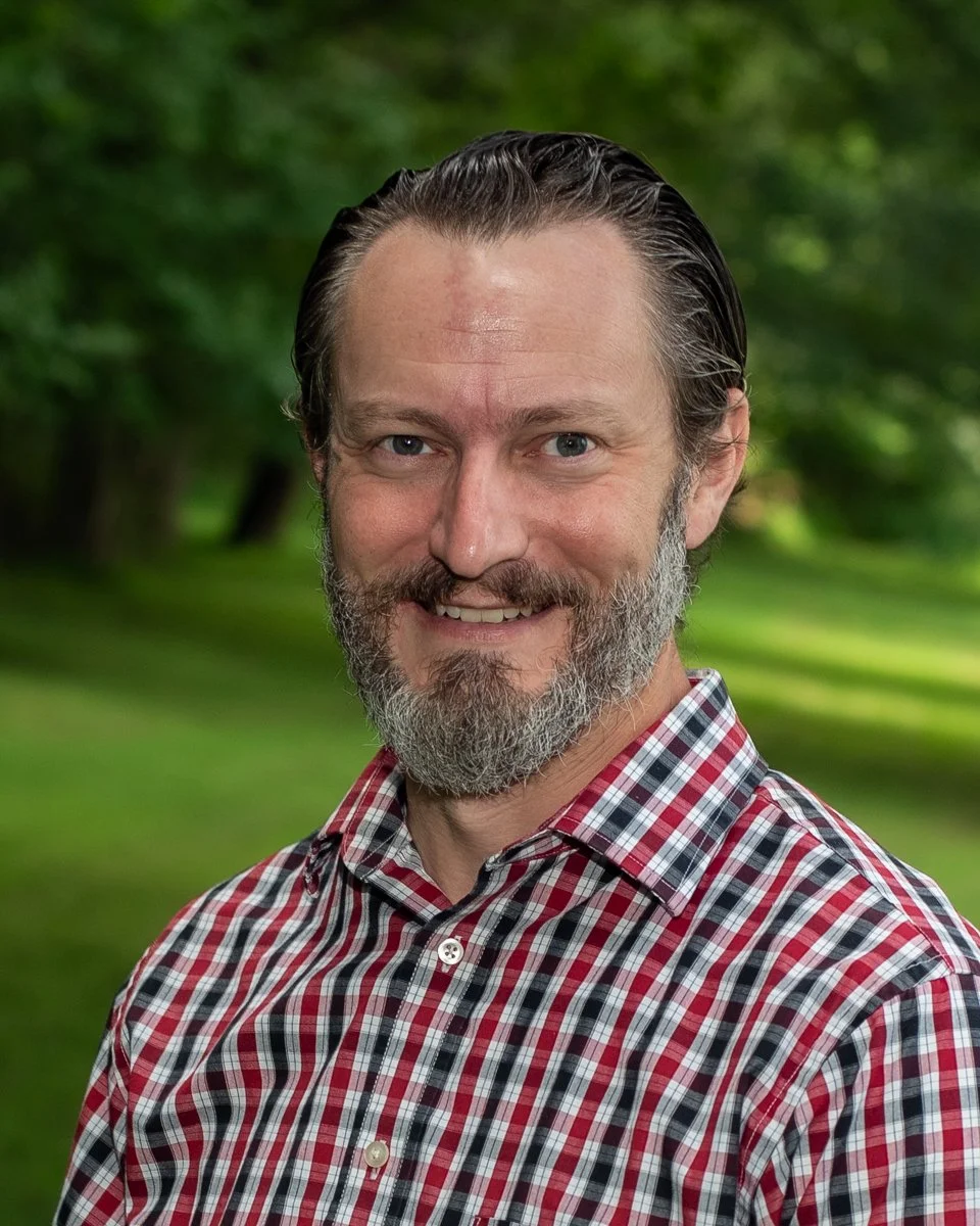 A middle-aged man with a beard and slicked-back hair is smiling outdoors, wearing a red, white, and black plaid shirt, with a green, blurred forest background.