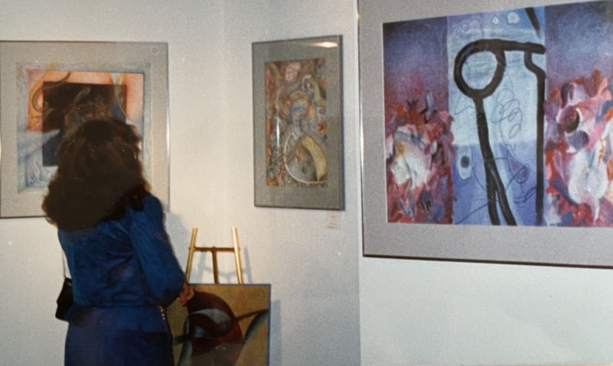 A woman in a blue jacket observing framed abstract artwork in a gallery, with paintings on the white walls behind her.