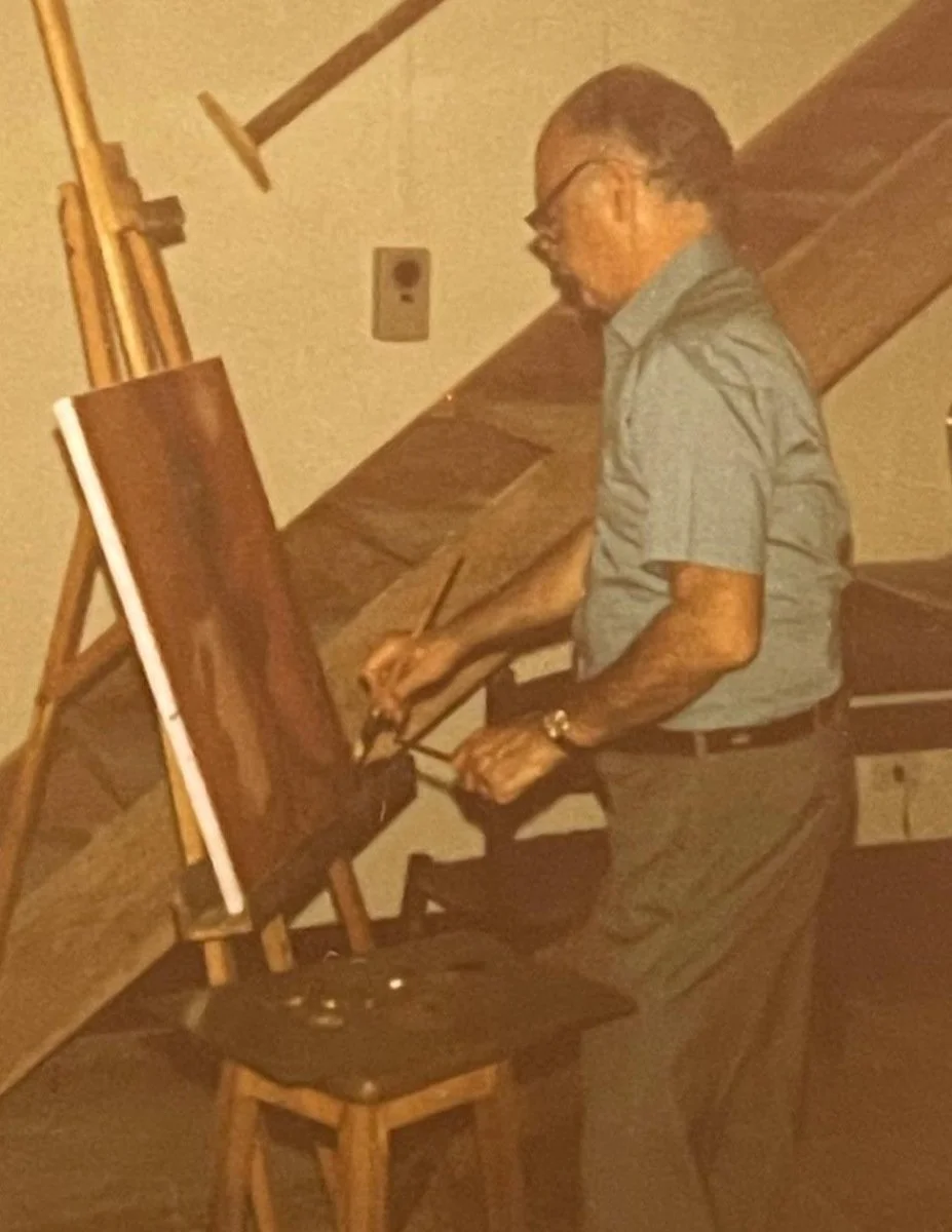 A man wearing glasses, a gray shirt, and dark pants is painting a landscape on a canvas using brushes in a room with wooden beams on the ceiling.