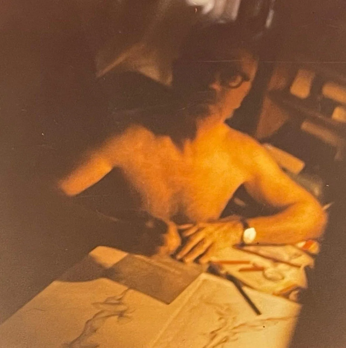 A shirtless man with glasses sitting at a table with papers and notebooks, and a dog with a light-colored coat resting on his shoulder in a dimly lit room.